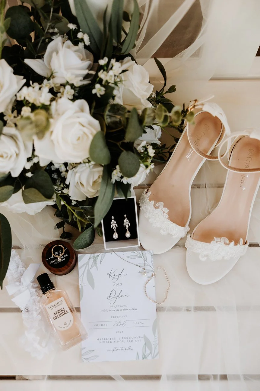 Bridal accessories and wedding items including white lace heels, a bouquet of white roses and greenery, pearl earrings, a delicate necklace, a wedding invitation, a perfume bottle, and a small container with a handwritten name on it.