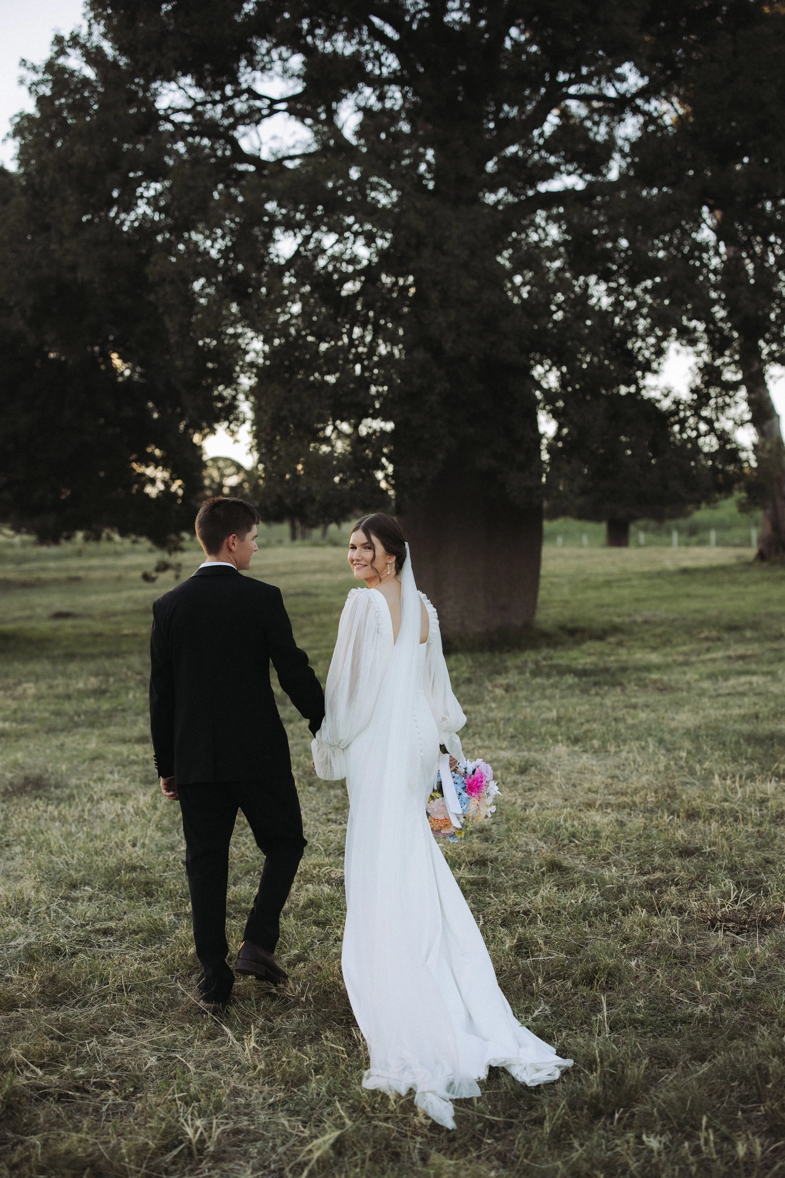A bride and groom walking hand in hand outdoors in a grassy area with large trees in the background, during the daytime.