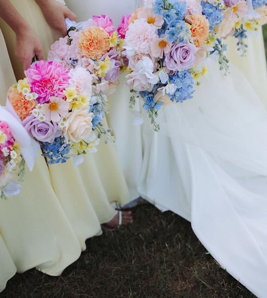 People holding bouquets of pink, purple, yellow, and blue flowers during a wedding or celebration outdoors.