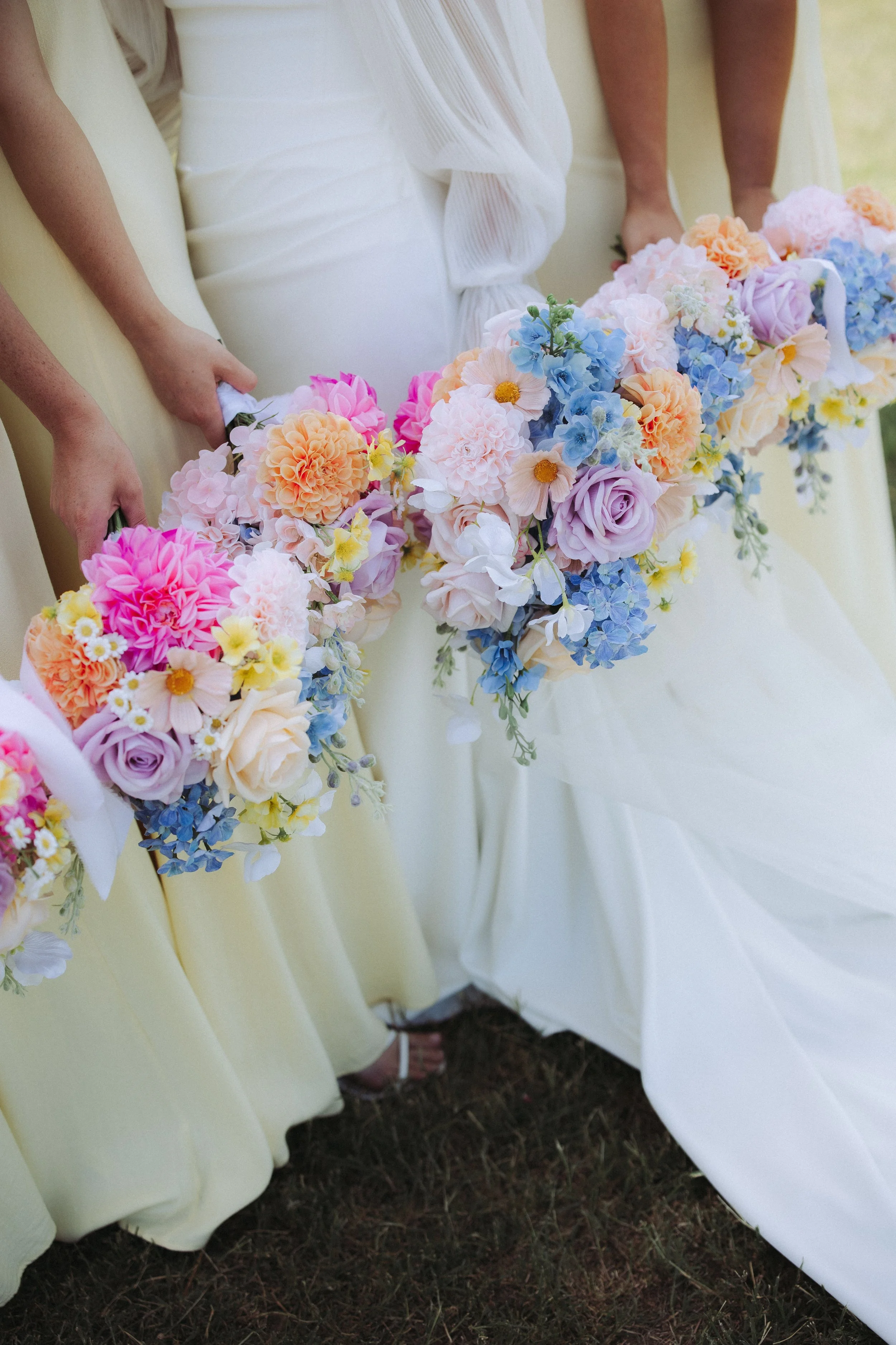 People holding floral bouquets at a wedding, standing on grass with their dresses touching the ground.