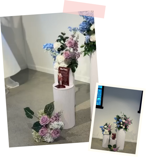 Flower arrangement with purple, pink, and white roses and greenery in a white vase, with a smaller matching arrangement on the floor, on a gray pedestal. A candle and card are inside the vase, set against a white wall with a black window.