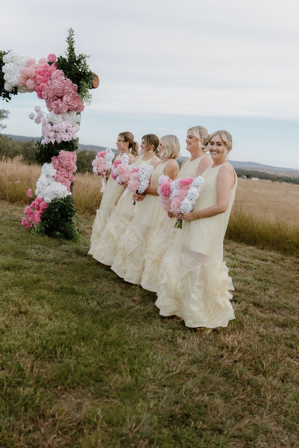 Six women in cream-colored dresses holding pink and white flower bouquets, standing outdoors next to a floral wedding arch in a field with a scenic landscape in the background.