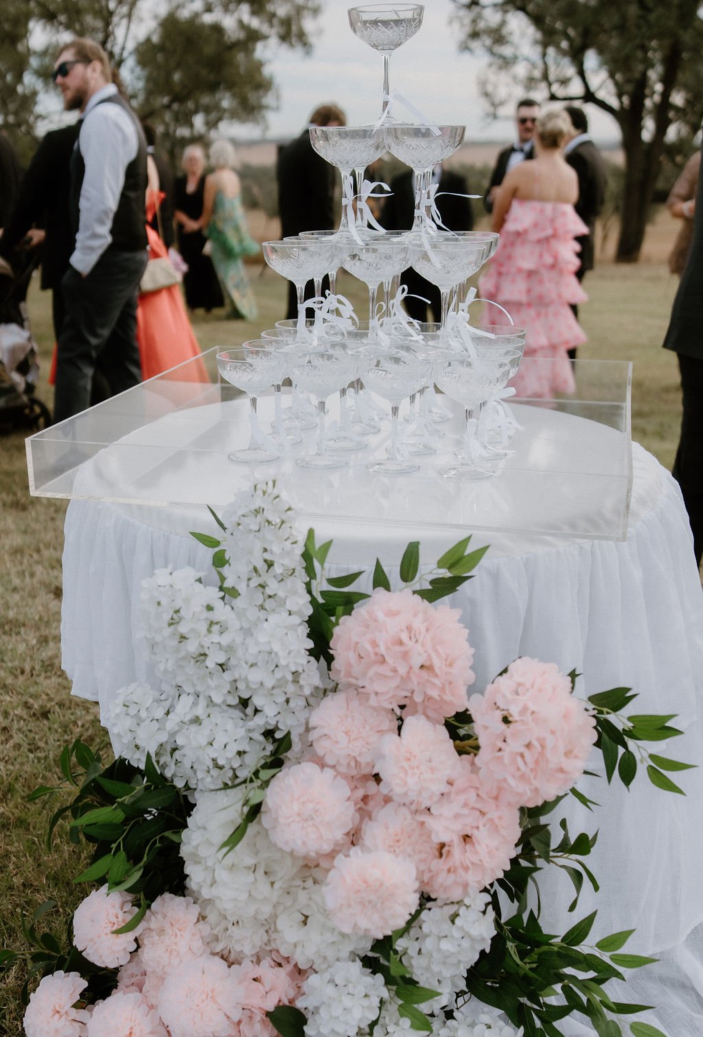 Empty champagne glasses stacked in a pyramid on a white table, decorated with pink and white flowers, during an outdoor event with people in formal attire in the background.