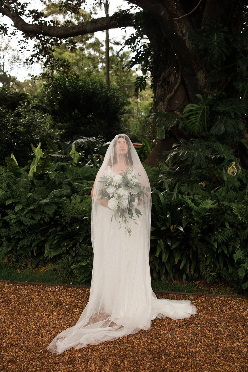 A bride in a white wedding dress holding a bouquet of white roses and greenery, standing outdoors in a lush, green garden with large trees and dense foliage, covered by a sheer veil over her face.