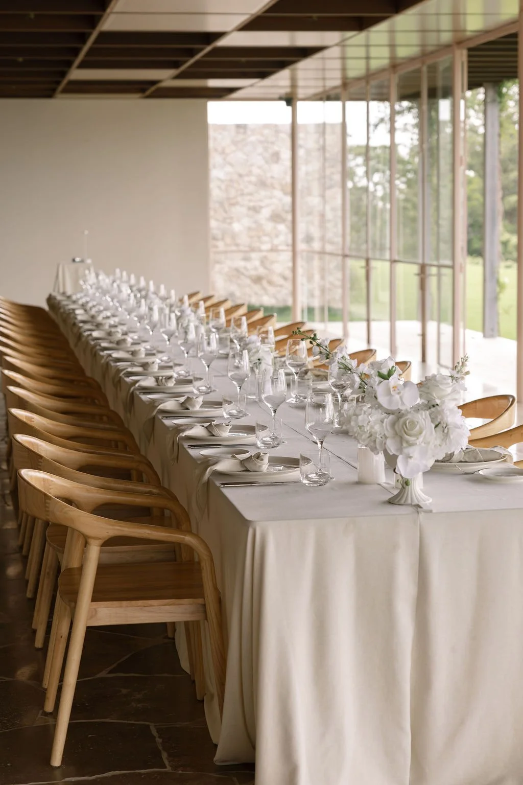 Elegant banquet table set with white tablecloth, floral centerpieces, glassware, and napkins in a bright, modern venue with large windows.