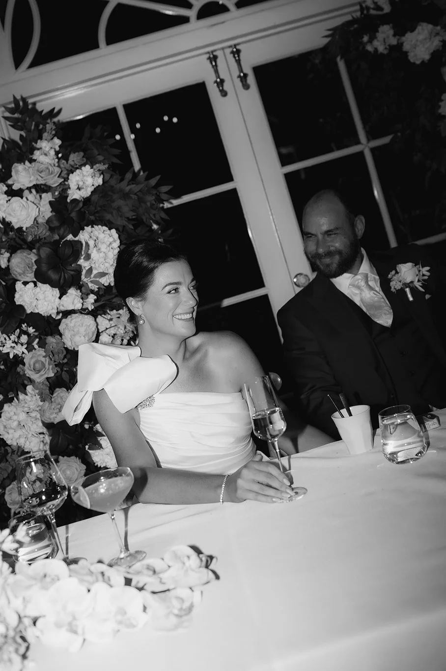 Bride and groom sitting at a wedding reception table with floral decorations, smiling and enjoying champagne.