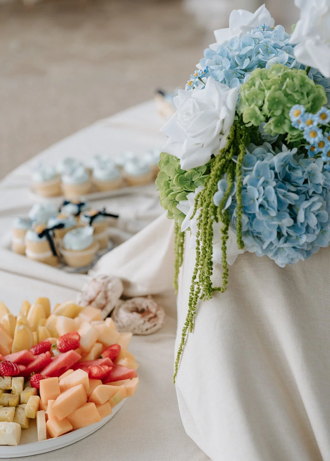 A table with a floral centerpiece of white, blue, and green flowers, with a tray of assorted cheeses and fruit, and small cupcakes in the background.