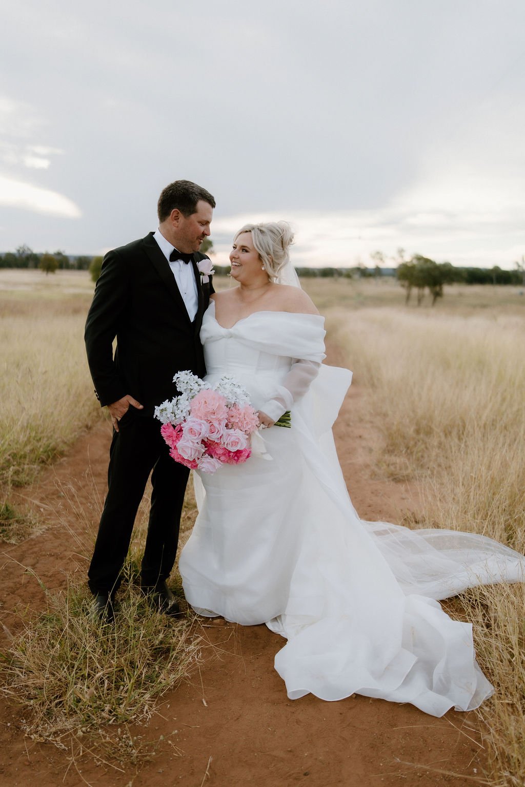 Bride in a white wedding gown holding a pink and white bouquet, standing next to a groom in a black tuxedo on a dirt pathway in a grassy field, with a cloudy sky in the background.
