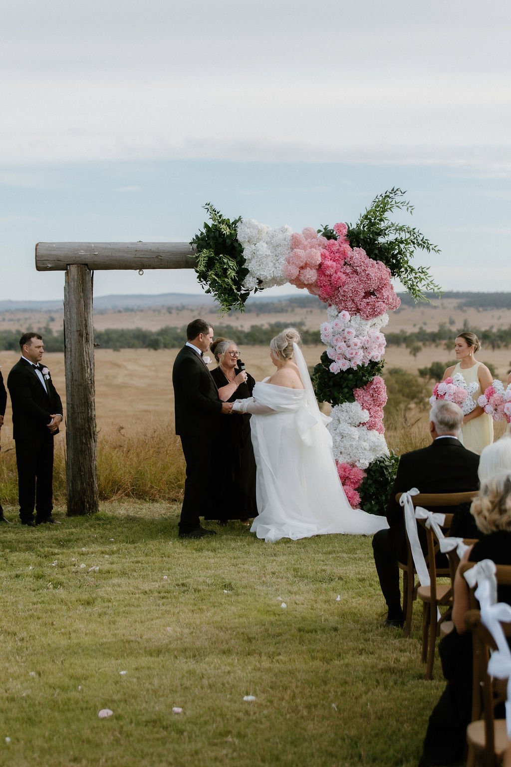 A wedding ceremony outdoors with a couple exchanging vows during sunset, under a wooden arch decorated with pink and white flowers, in a field with guests seated on wooden chairs.