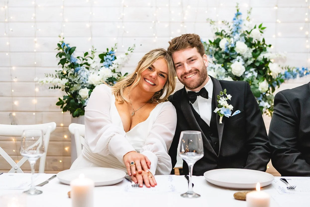 Bride and groom smiling at wedding reception, sitting at table decorated with candles and floral arrangements in the background.