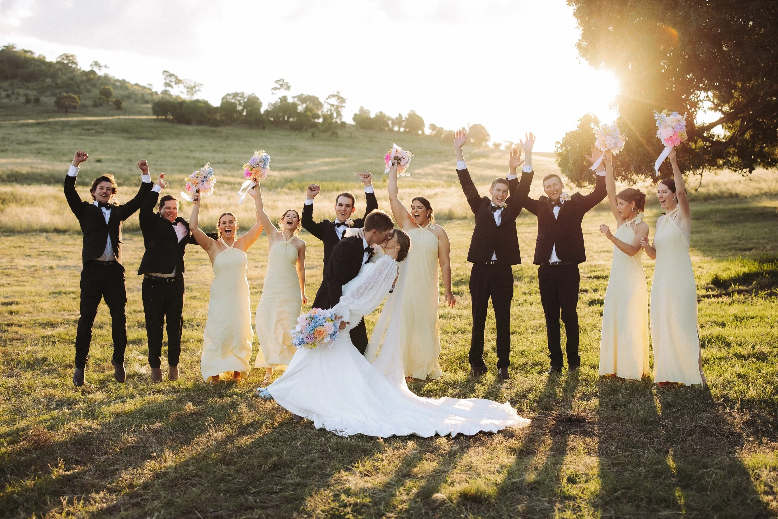 A group of people dressed in wedding attire celebrating outdoors during sunset, with some holding bouquets and others raising their hands.