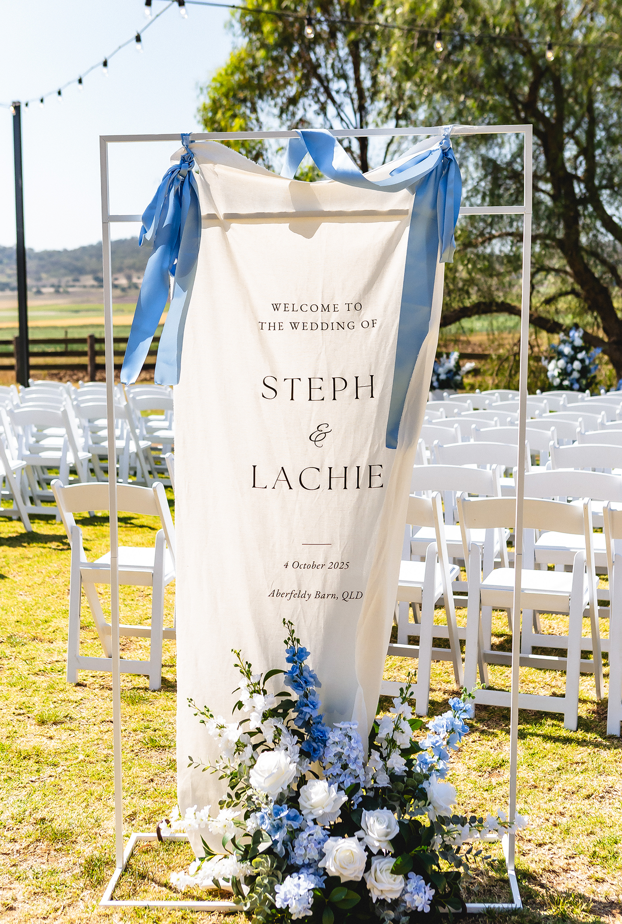 An outdoor wedding ceremony setup with a decorated welcome banner for the wedding of Steph and Lachie, featuring white and blue flowers and chairs arranged under string lights.