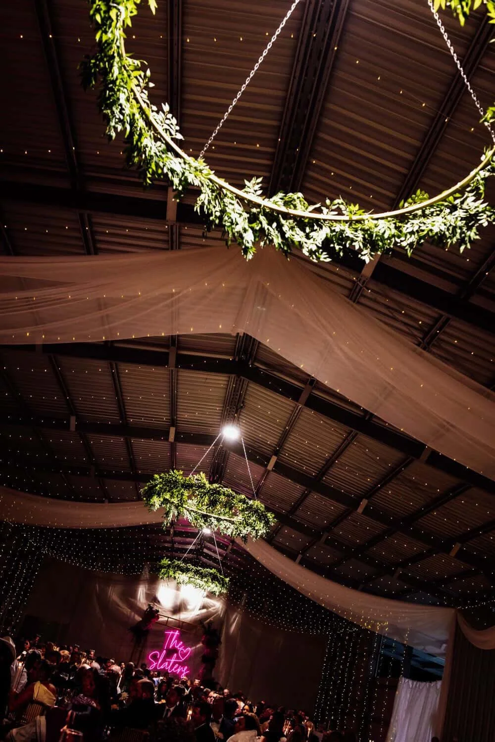 Decorative event space with hanging greenery and lights, a stage with pink neon signs reading 'The Slater', and a crowded audience.
