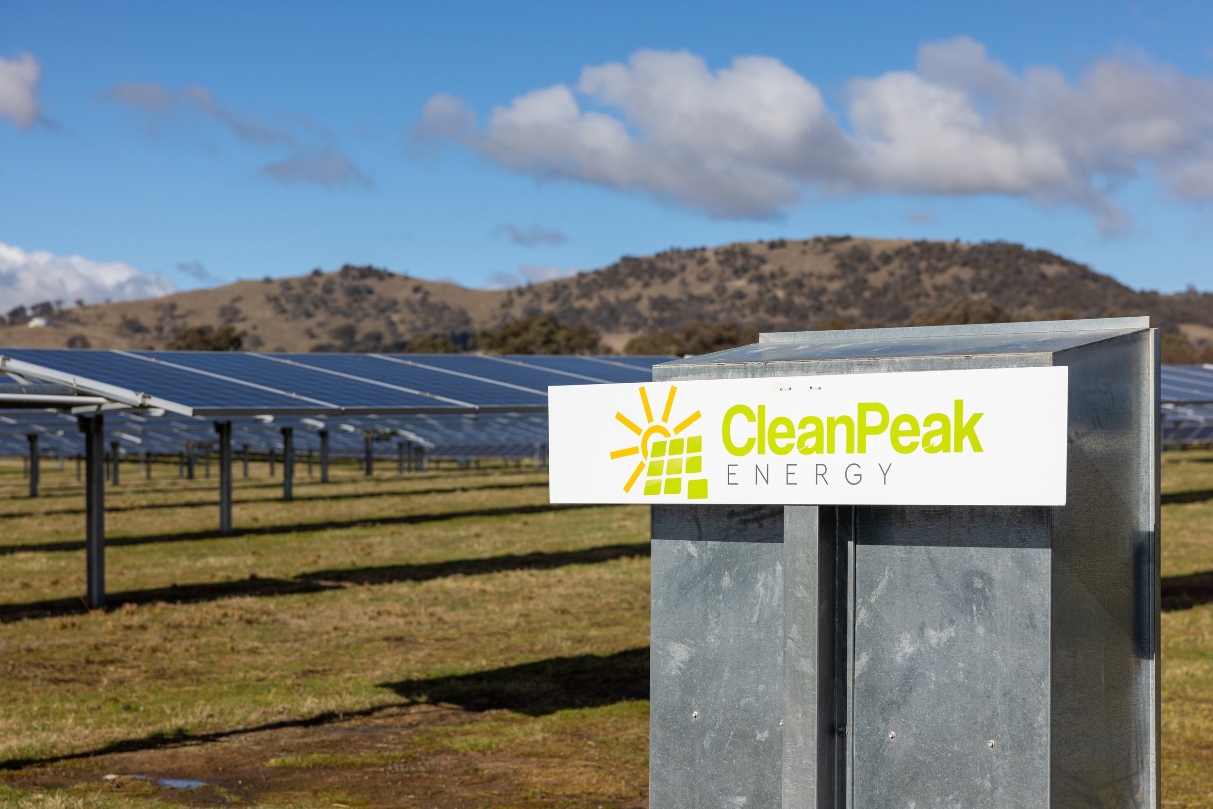 A solar panel farm with a sign that reads 'CleanPeak Energy' in front of the panels, with mountains and a partly cloudy sky in the background.