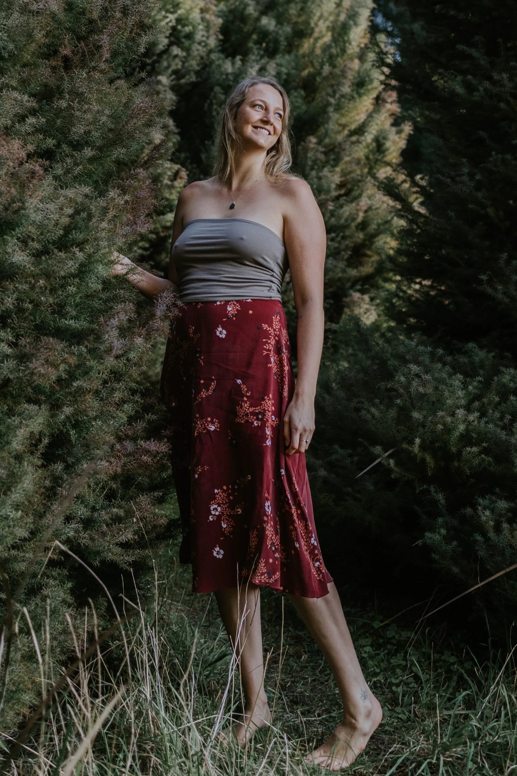 A woman standing outdoors surrounded by greenery and trees, smiling and looking away from the camera, wearing a strapless top and a red floral skirt.