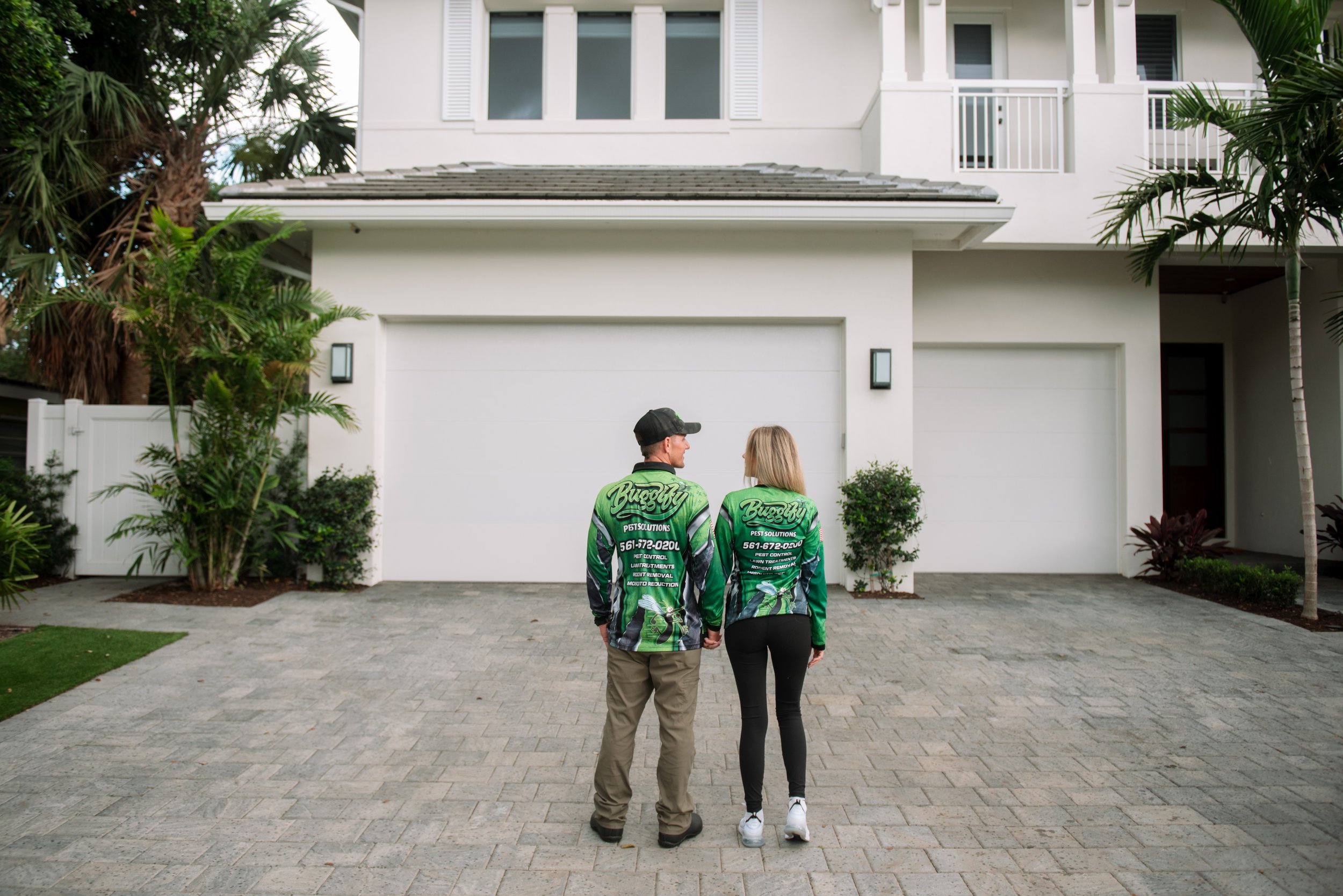 Two people walking away from the camera toward a white modern house with two floors, a garage door, and palm trees on the front yard, wearing green pest control uniforms.
