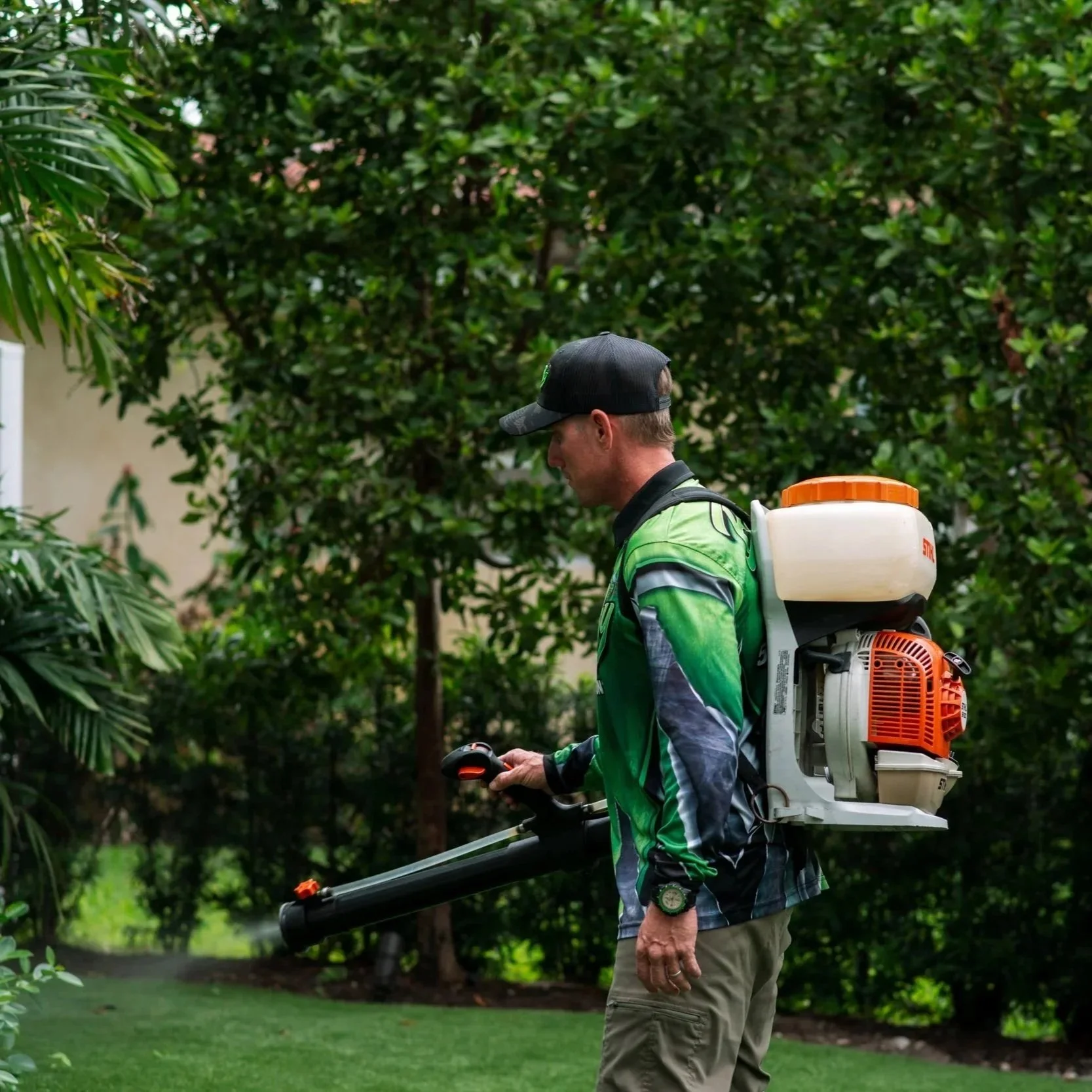 A man wearing a black cap and green jacket is trimming bushes with a hedge trimmer in a backyard with lush green trees and grass.
