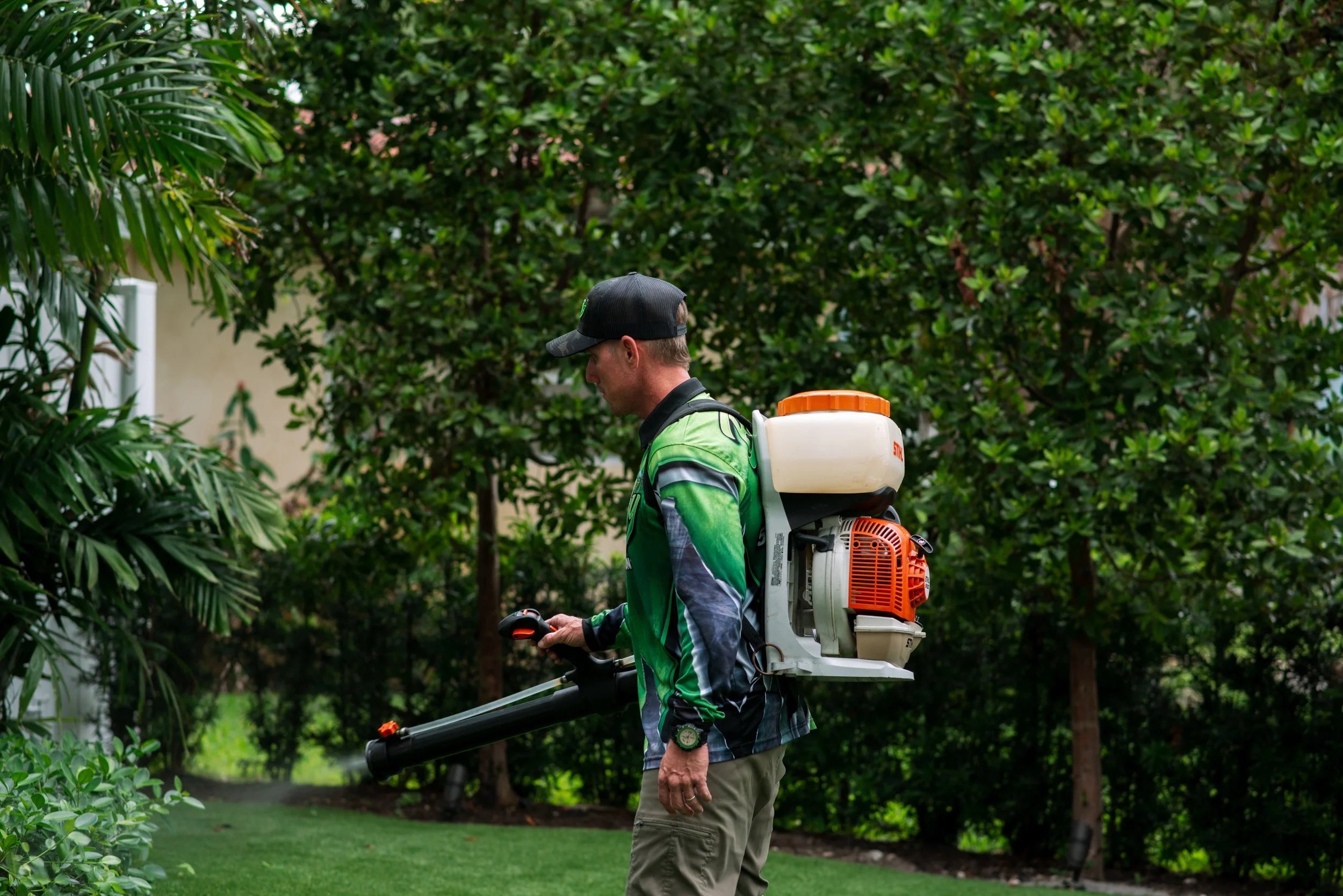 A man wearing a black cap, green jacket, and khaki pants using a leaf blower in a garden surrounded by green trees and bushes.