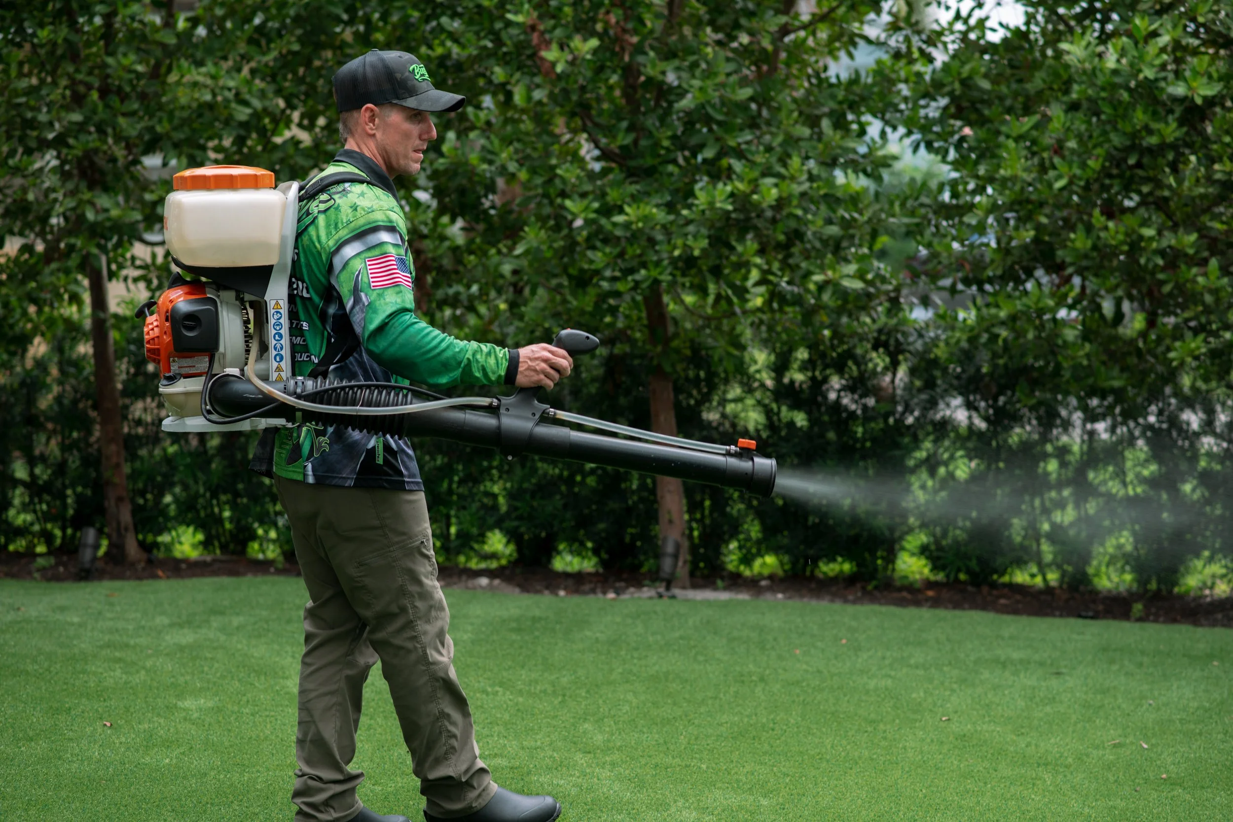 A man wearing a black cap and a green sports jersey is using a backpack sprayer to spray the grass in a backyard with trees and green lawn.