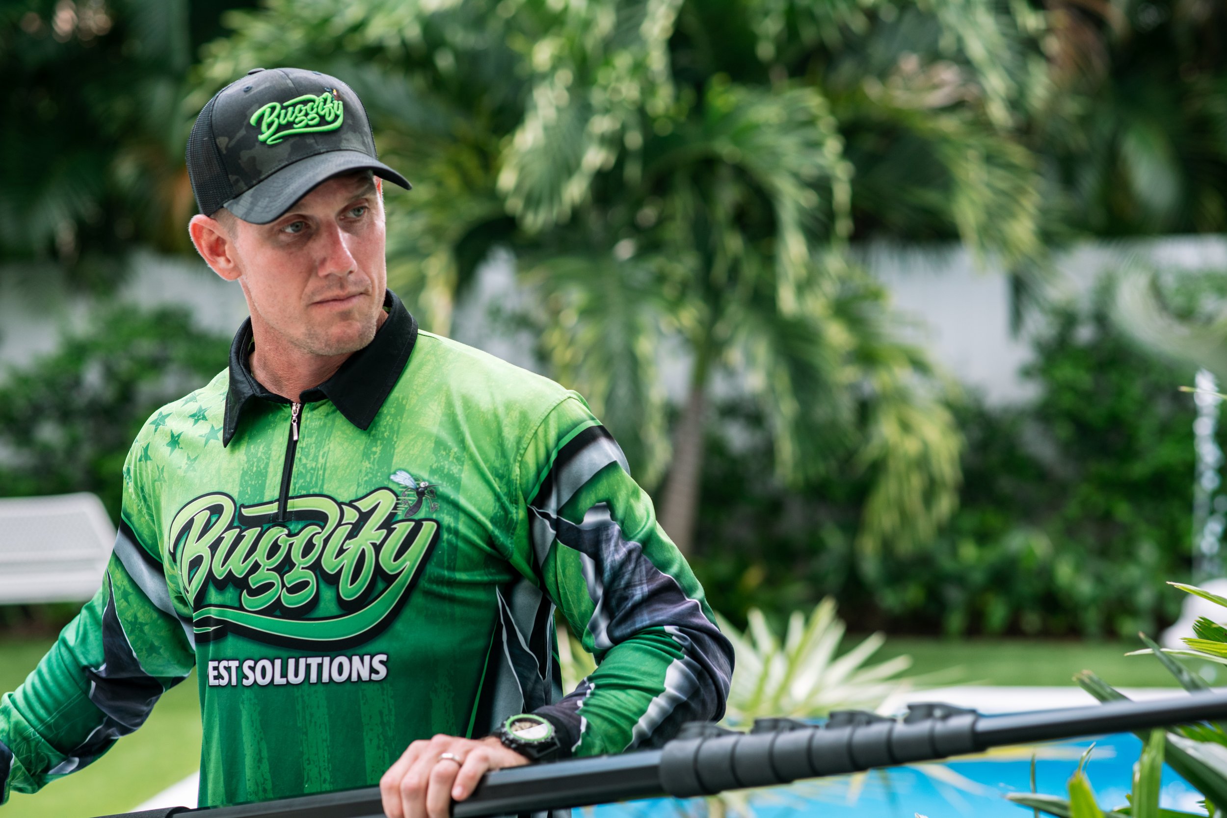 A man wearing a green and black sports jersey with the word "Buggy" on it and a matching cap with the same logo. He is outdoors with lush greenery in the background, and he appears to be concentrating on something in front of him.
