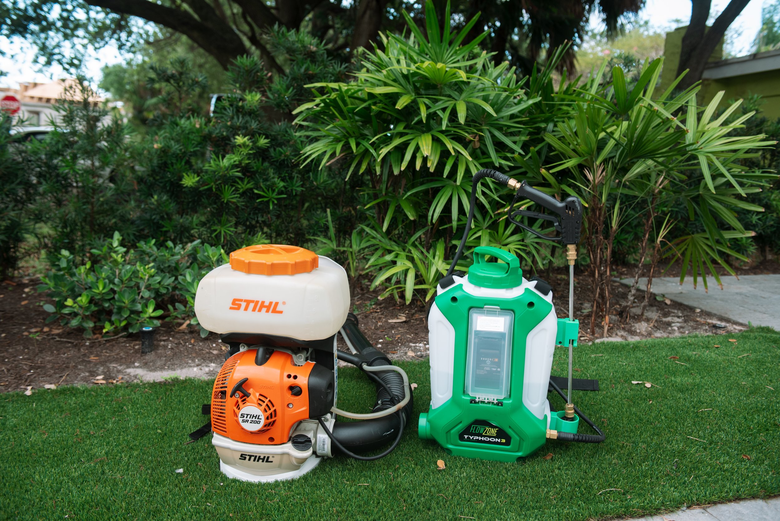 Lush green plants and bushes in a garden with gardening equipment including a leaf blower, sprayer, and weed sprayer in the foreground.