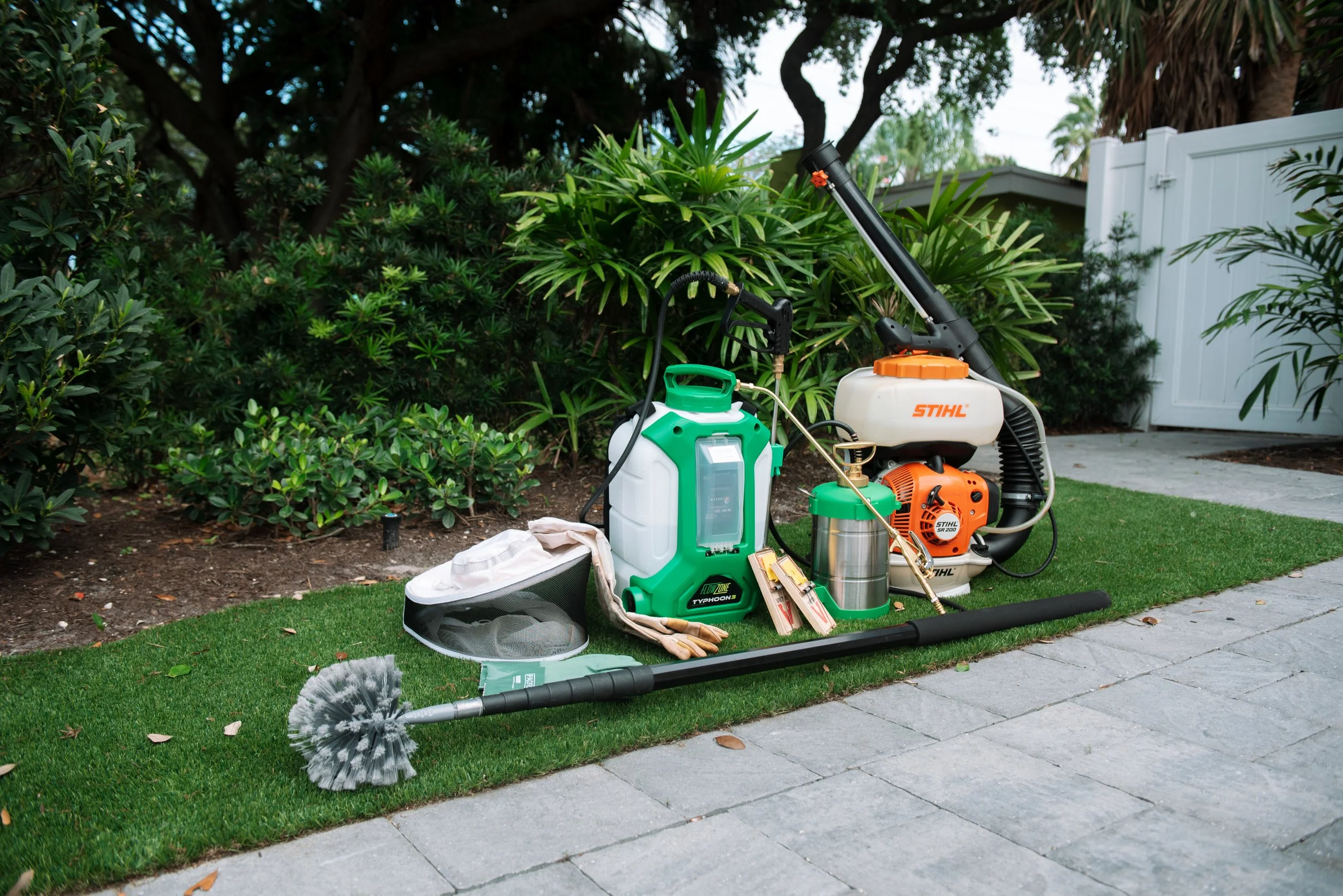 Lawn care tools on a lawn, including a leaf blower, sprayer, garden gloves, and a leaf rake, with shrubs, trees, and a white fence in the background.