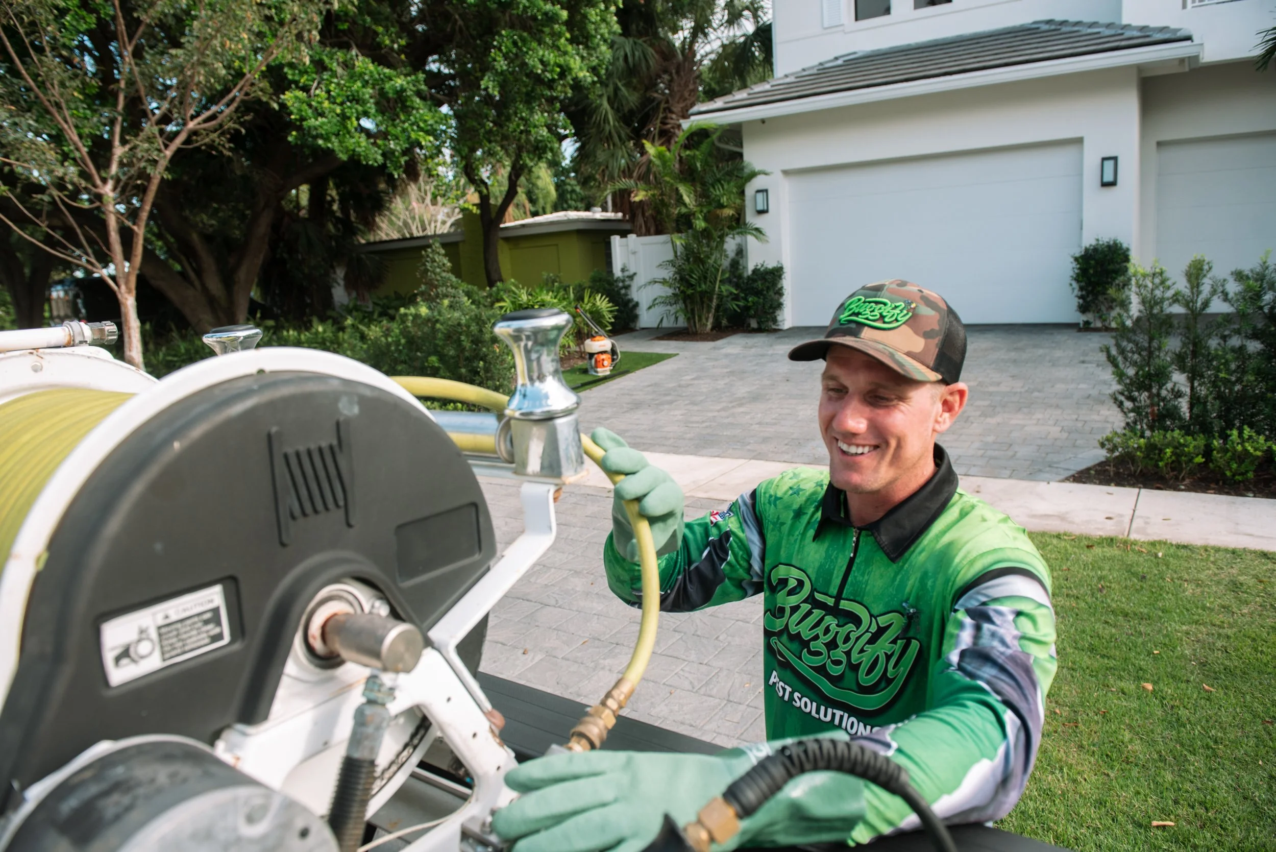 A smiling man wearing a green and black shirt and a camouflage cap is working on outdoor equipment near a driveway with a white house and trees in the background.