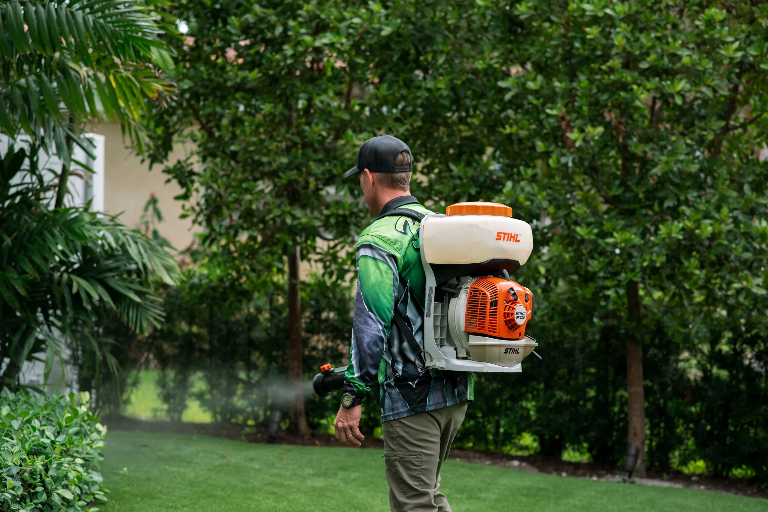 A man wearing a green sports jersey, khaki pants, and a black cap is walking in a lush green garden with a backpack sprayer on his back, spraying plants.