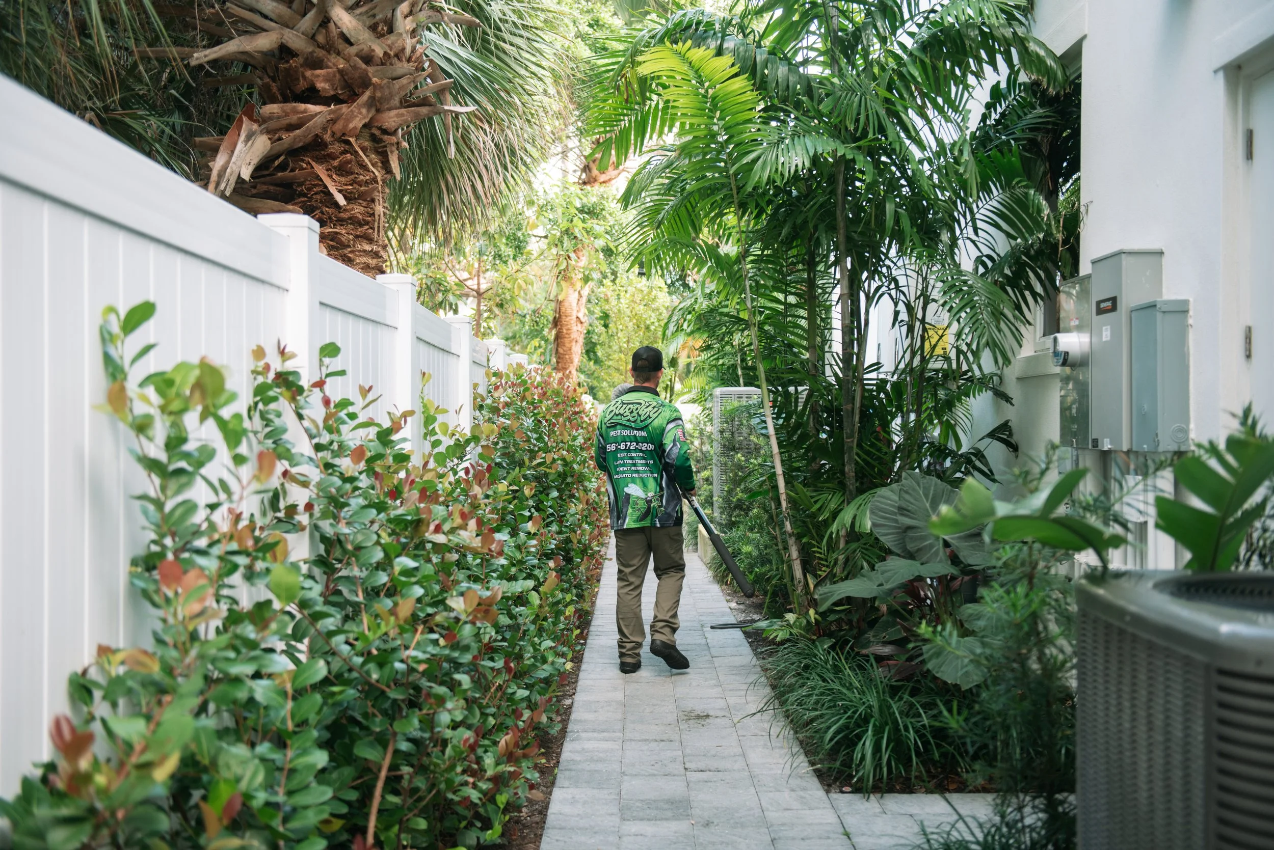 A person wearing a green uniform with pest control branding walks along a narrow sidewalk bordered by lush tropical plants and a white fence, with utility boxes on the wall to the right.