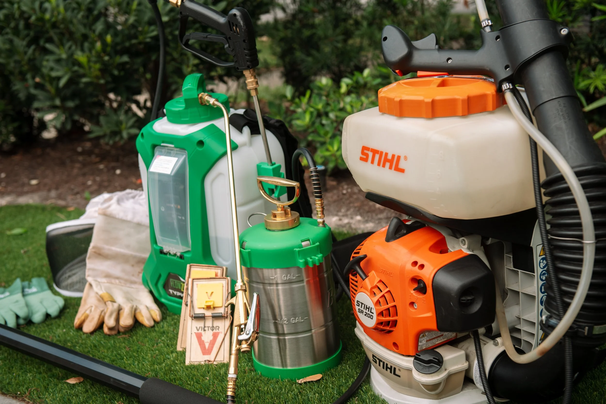 Lawn or garden tools and equipment, including a green and white tank sprayer, orange and white chainsaw, and various accessories, arranged outdoors on a black tray with a grassy and shrub background.