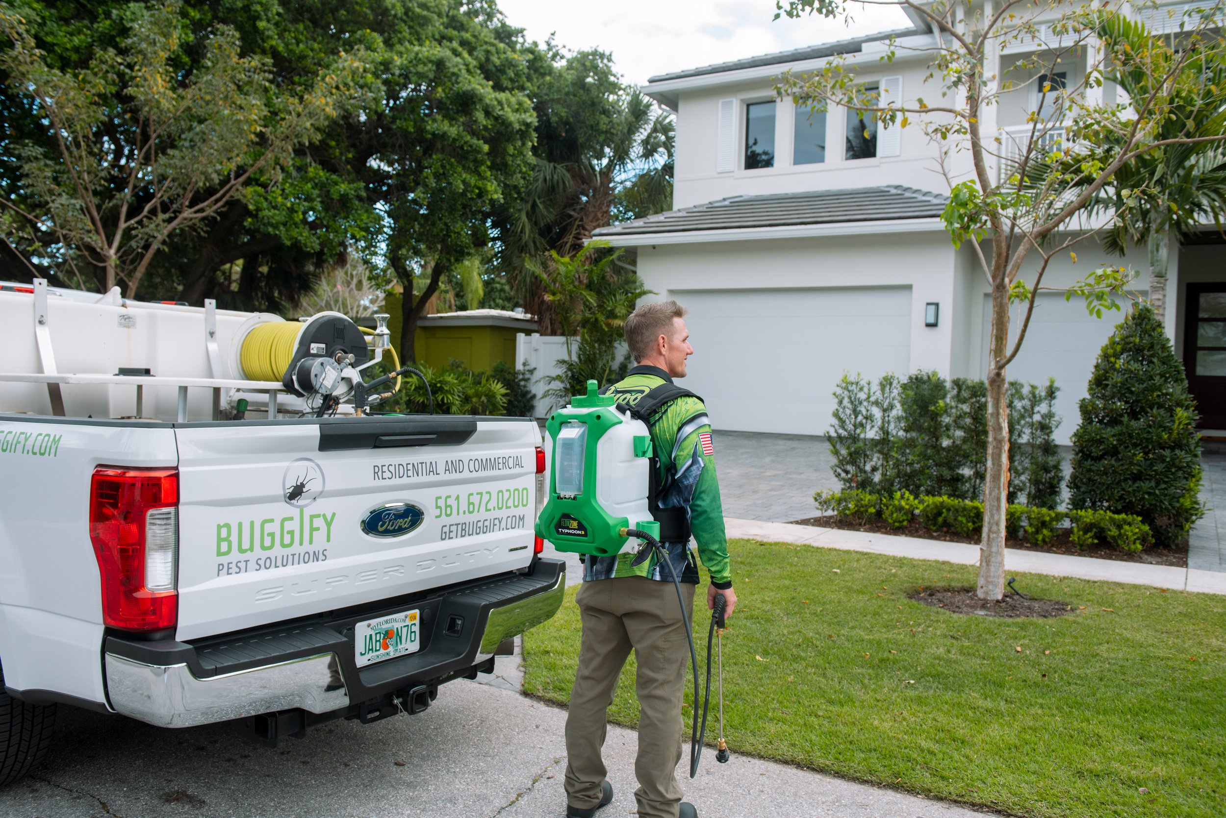 A pest control worker in a green uniform with an insecticide sprayer stands next to a white pest control truck parked in front of a modern white house with a driveway, trees, and lush green lawn.