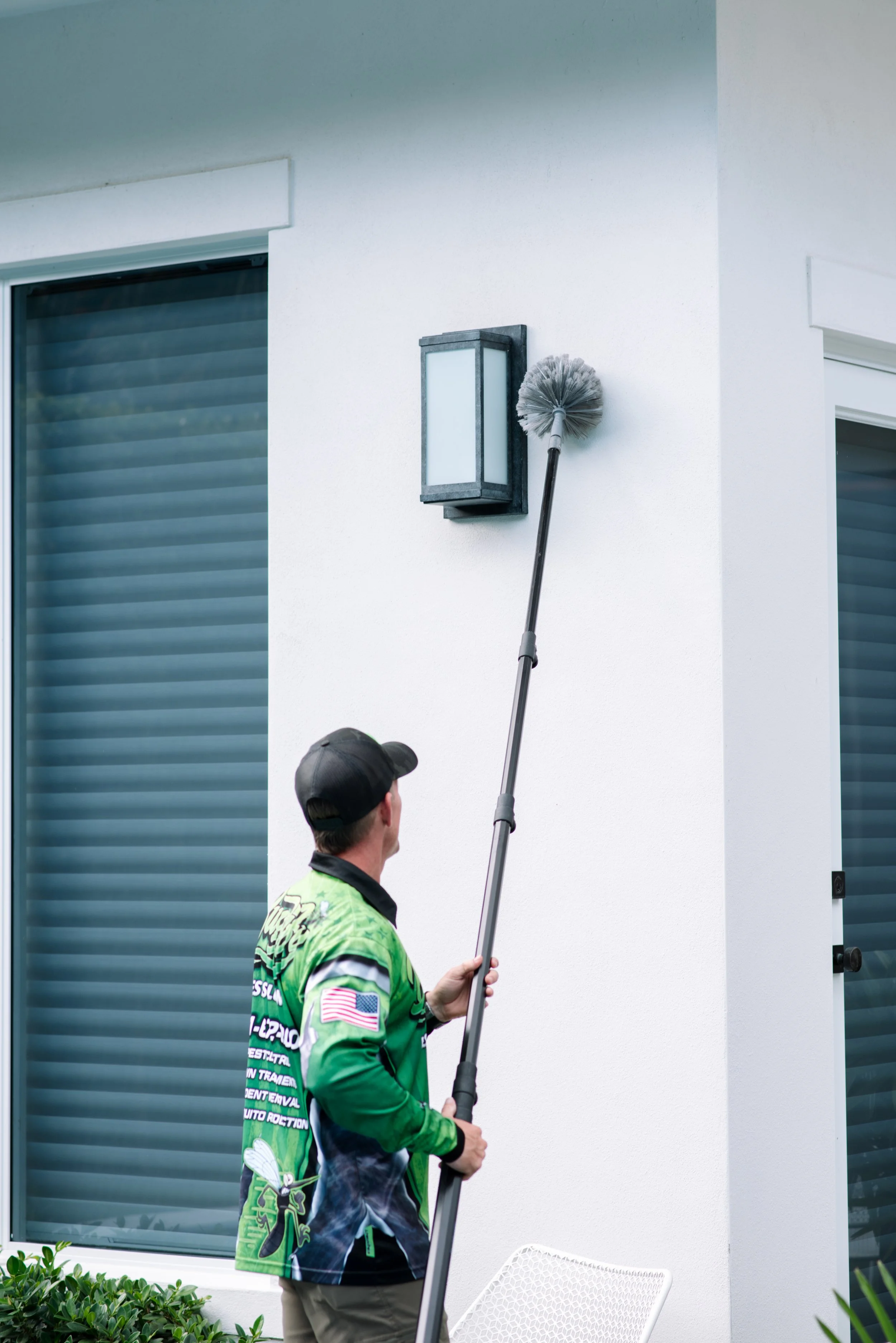 A person wearing a green racing shirt and a black cap is standing outside near a white wall while using a long-handled cleaning tool to clean a light fixture attached to the wall. There are two windows with closed blinds and some green bushes in the foreground.