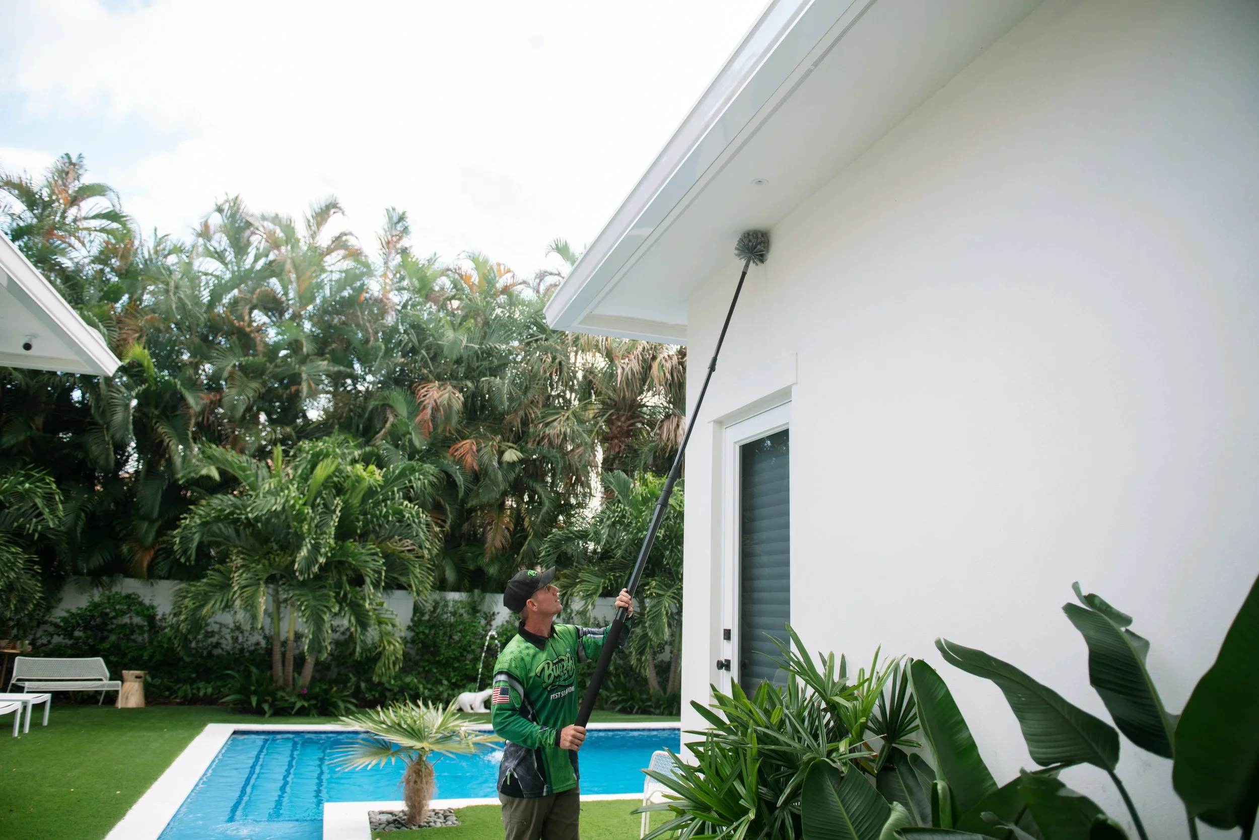 A person in a green and black jacket and black cap is using a pole tool to clean the upper part of a white building's exterior wall. The scene is set in a backyard with lush green trees, a swimming pool, and outdoor furniture.