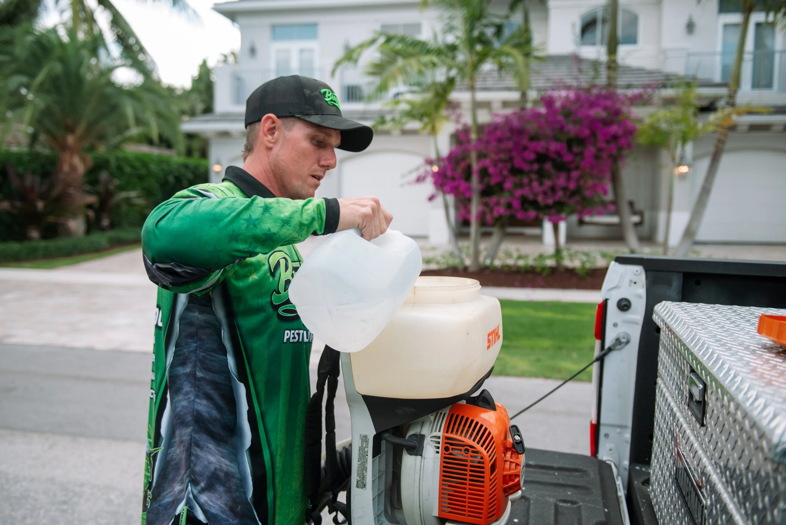 A man wearing a green pesticide suit and a black baseball cap operating a leaf blower on the back of a pickup truck, with a house and pink flowering bush in the background.