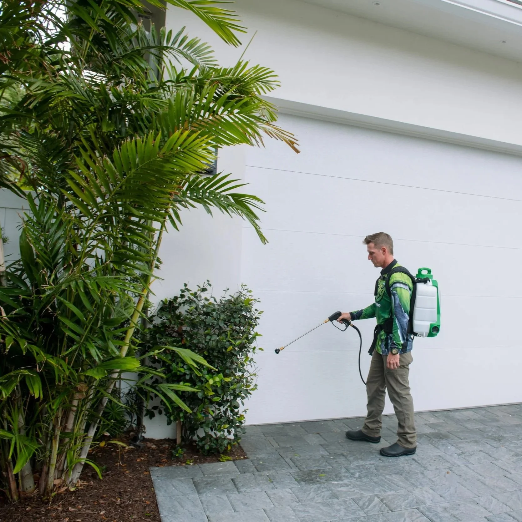 A man in a green outdoor jacket and khaki pants is using a sprayer on bushes next to a white building wall with a paved sidewalk.