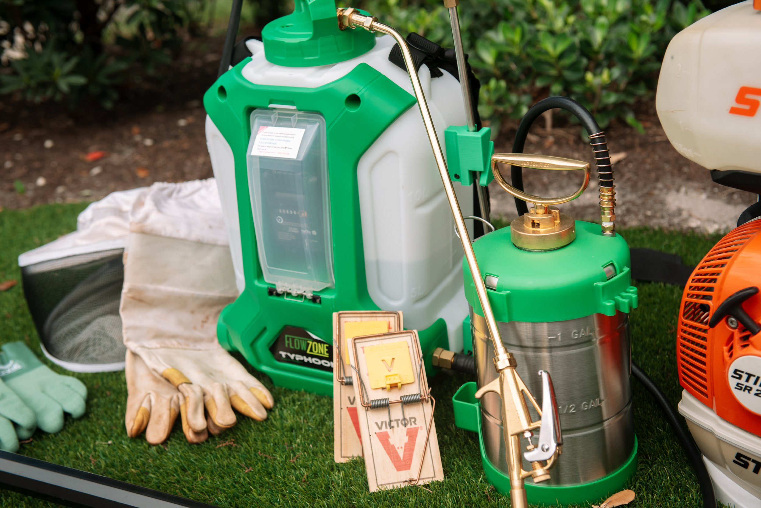 Green and white backpack sprayer with brass nozzle, hoses, and tanks, surrounded by work gloves, spray bottles, and other gardening equipment on grass.