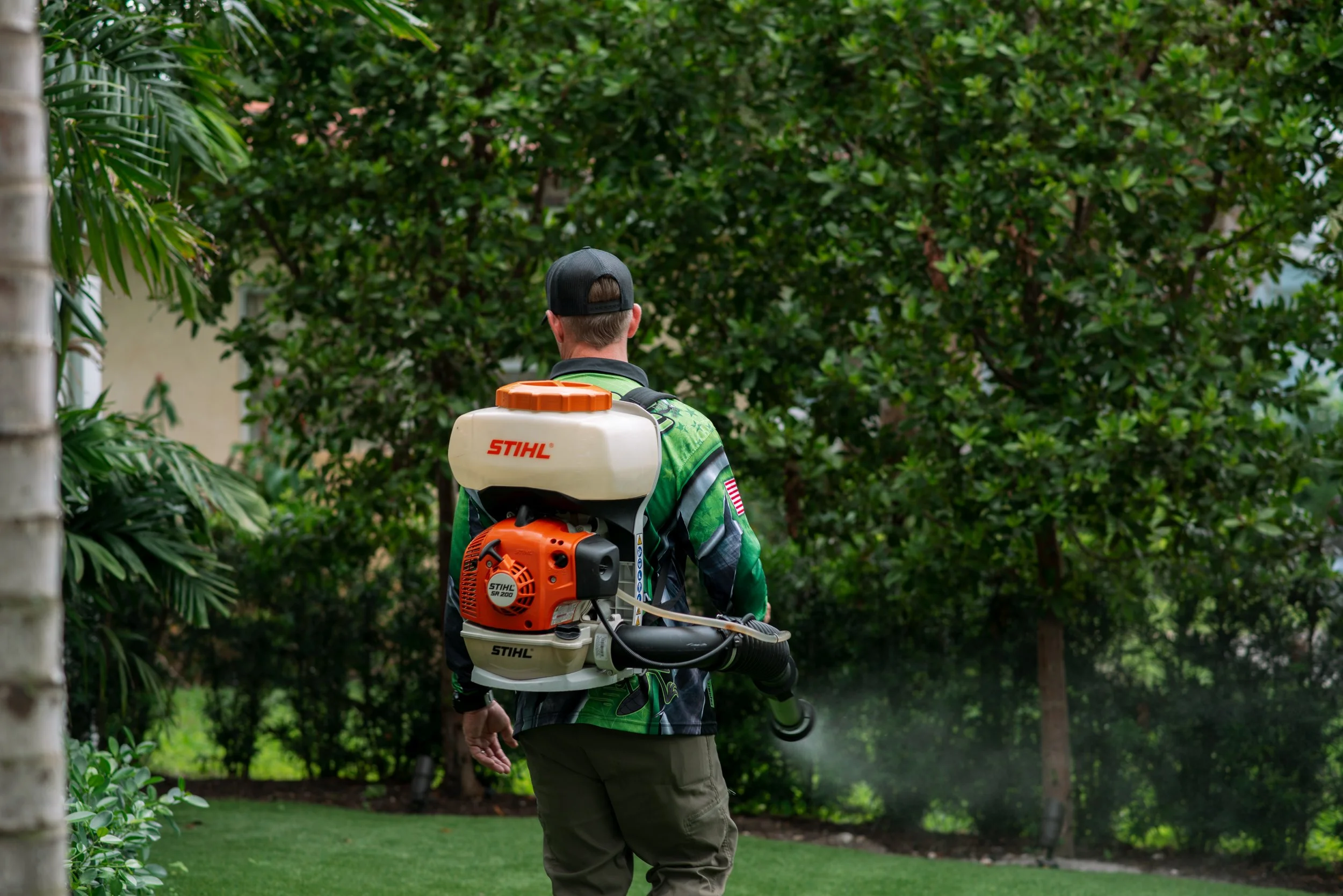 A man wearing a green jacket and black cap is using a STIHL backpack leaf blower in a backyard with green trees and bushes.