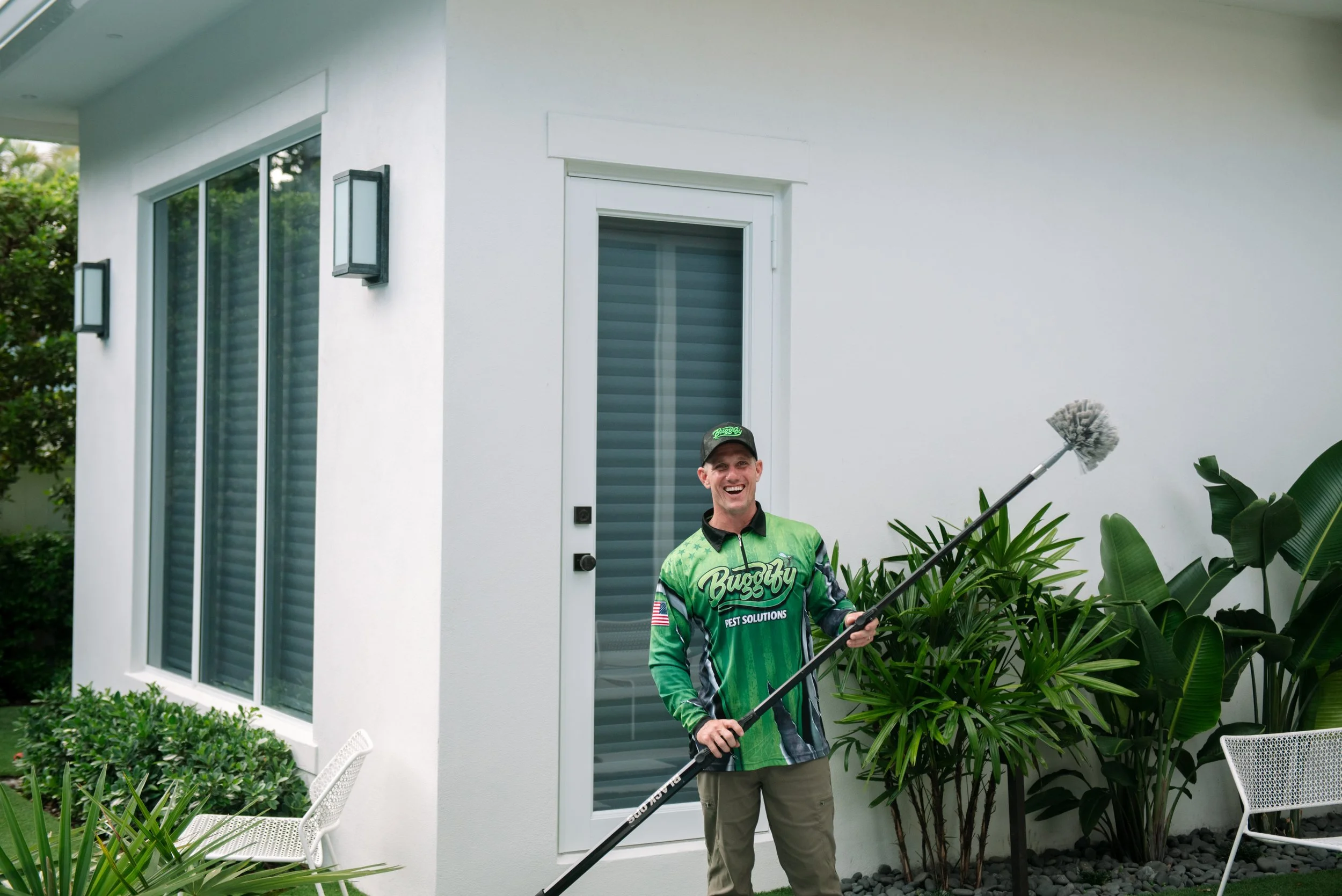 A pest control technician holding a sprayer outside a modern white house with large windows and lush green plants.