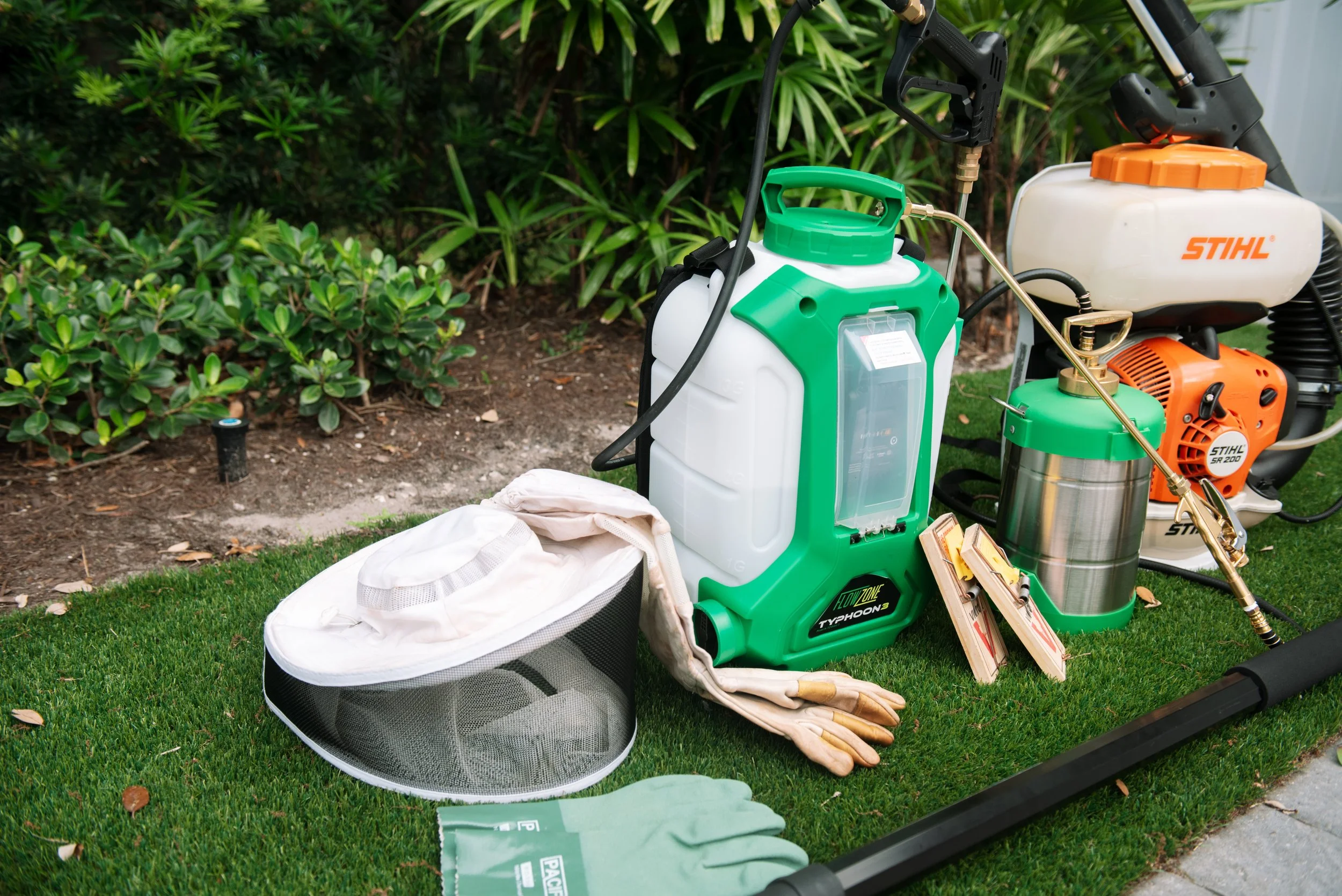 Lawn care equipment including a weed sprayer, leaf blower, and garden gloves arranged on a grass lawn with bushes in the background.