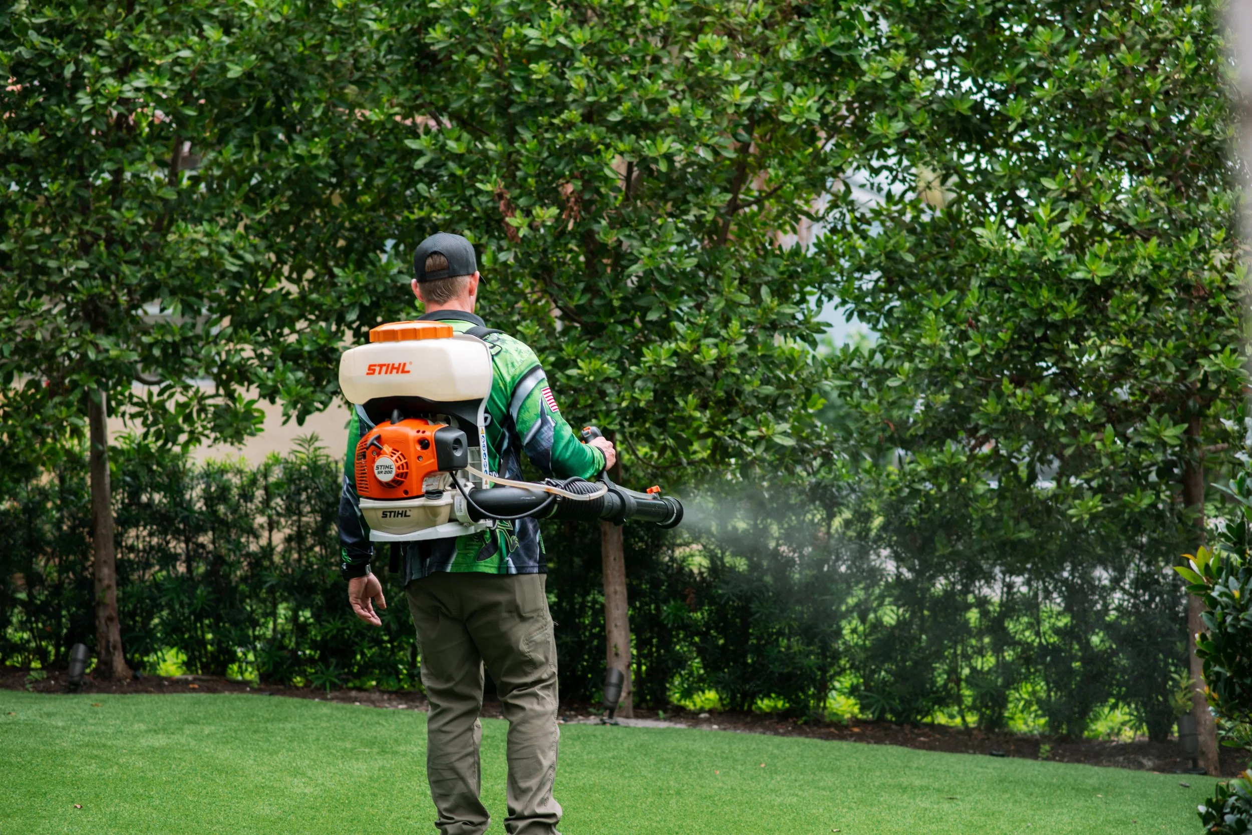 A person wearing a green jacket, khaki pants, and a black cap is using a leaf blower to clean leaves on a green lawn. The person has a backpack-style leaf blower with an orange and white engine labeled 'Stihl.' There are trees and bushes in the background.