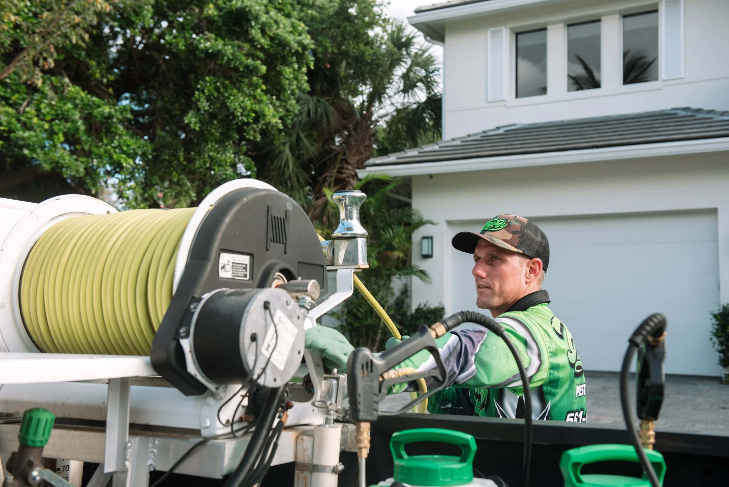A man wearing a camouflage cap and a reflective vest working on equipment with yellow hoses in front of a modern white house with large windows, surrounded by greenery.