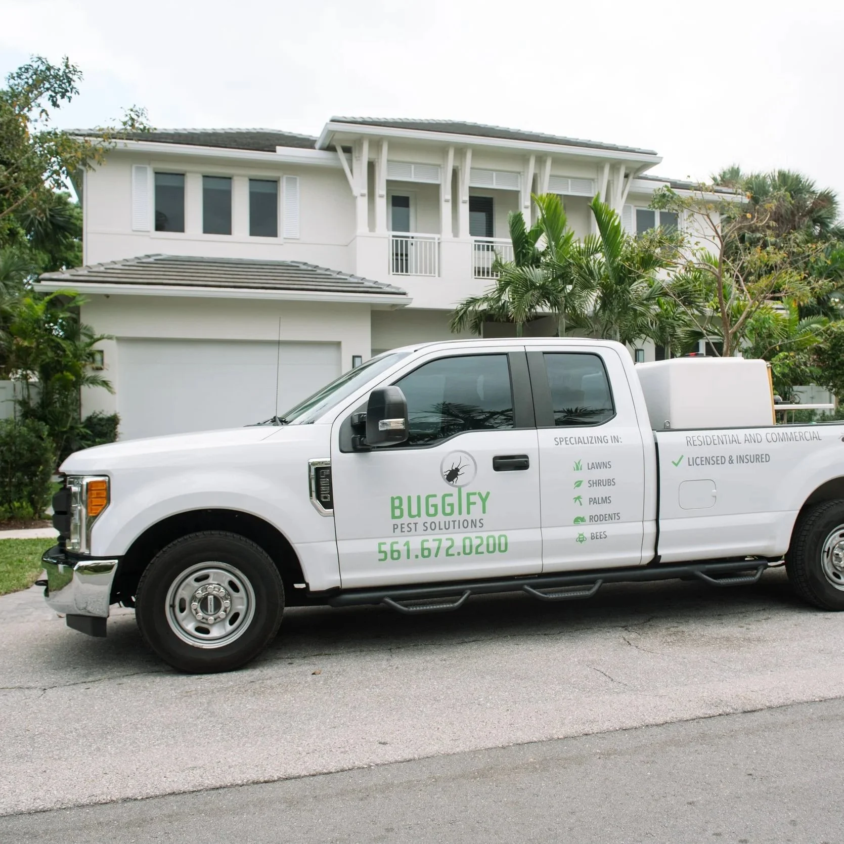 A white pest control service truck parked in front of a two-story white house with a balcony, surrounded by green trees and plants.