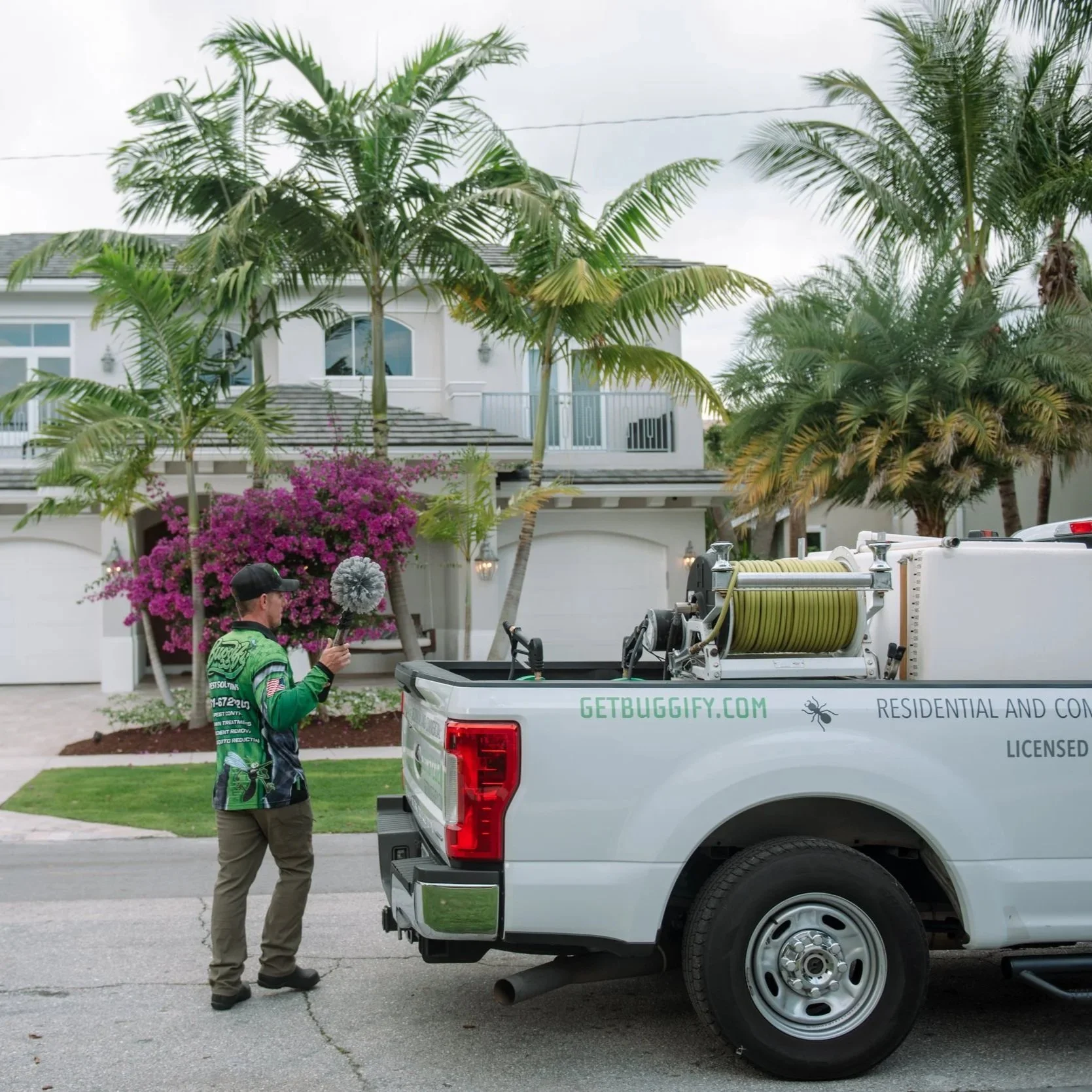 A man wearing a green shirt and khaki pants stands next to a white truck with yard equipment in its bed. In the background, there are palm trees, a purple flowering bush, and a white two-story house with balconies.