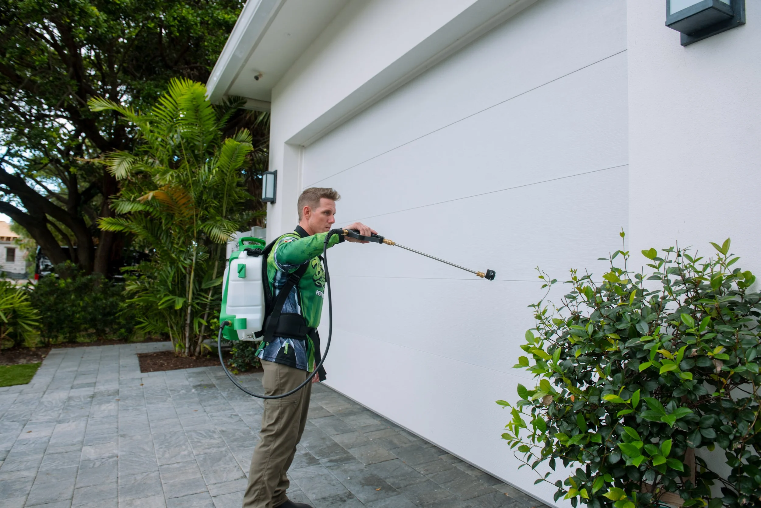 A man wearing a green and black jacket and khaki pants is spraying a white wall outside of a house with a garden spray. The house has a gray garage door, a black outdoor light, and is surrounded by green trees and bushes on a paved walkway.