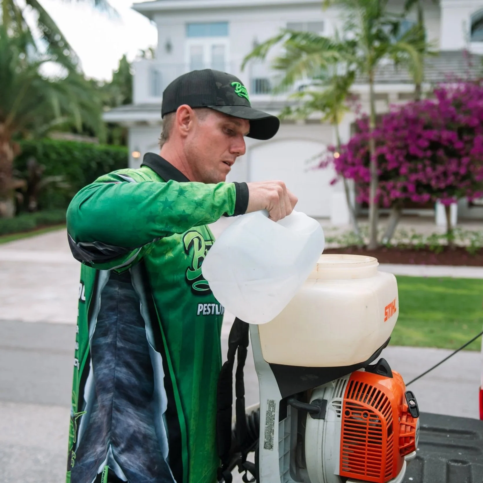 A man in a green and black sports jersey and black cap operating a backpack leaf blower outdoors. There is a residential house and purple flowering bush in the background.