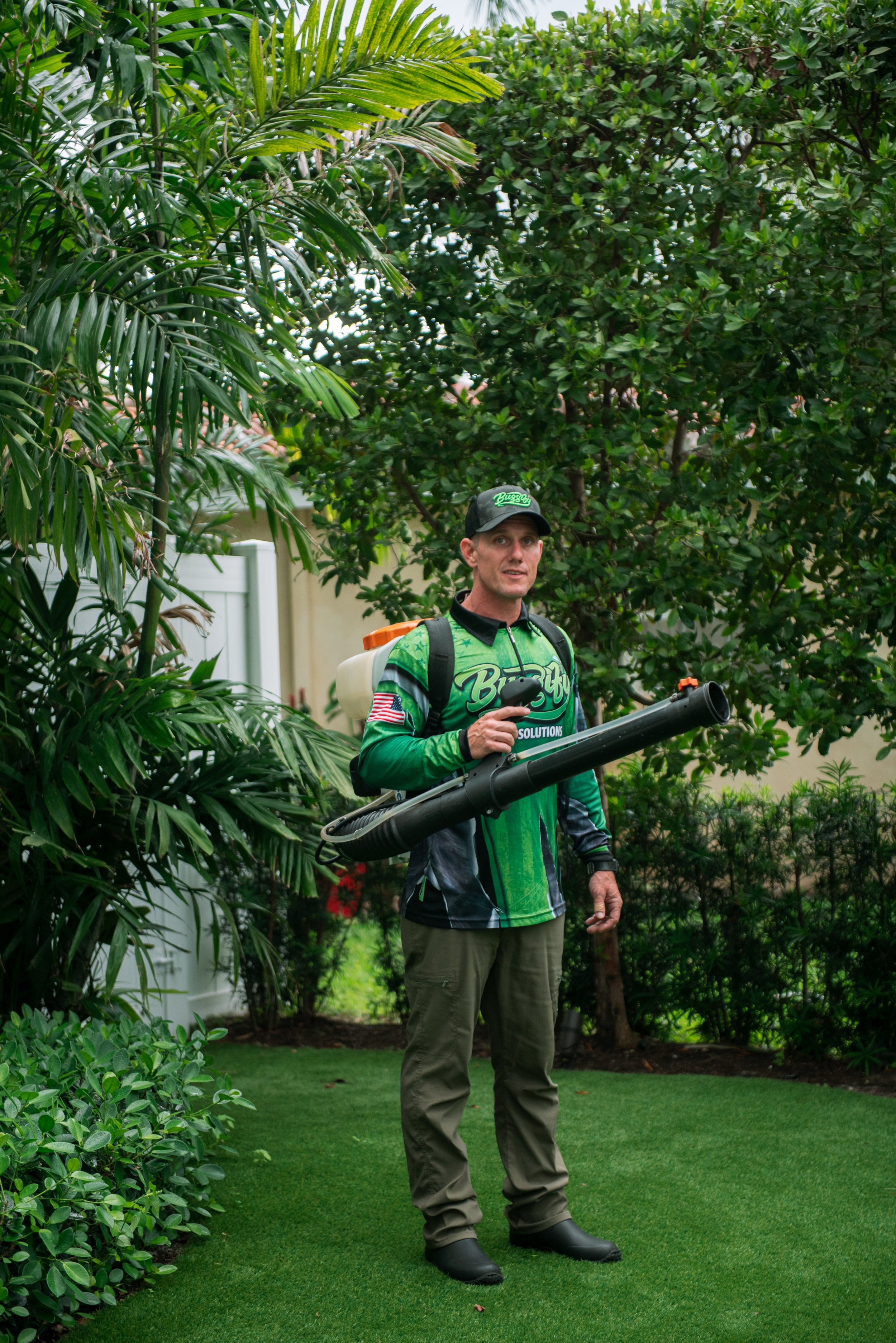 A man standing outdoors among lush green bushes and trees, holding a leaf blower in his right hand, wearing a black cap, a green and black gardening jacket, khaki pants, and black boots.