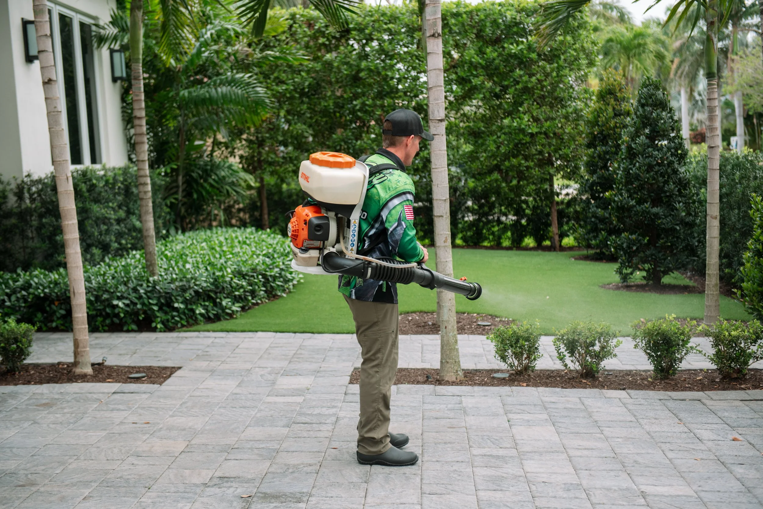 A man wearing a cap, green jacket with American flag patch, khaki pants, and black boots using a backpack leaf blower to clear leaves on a paved walkway surrounded by trees and bushes.