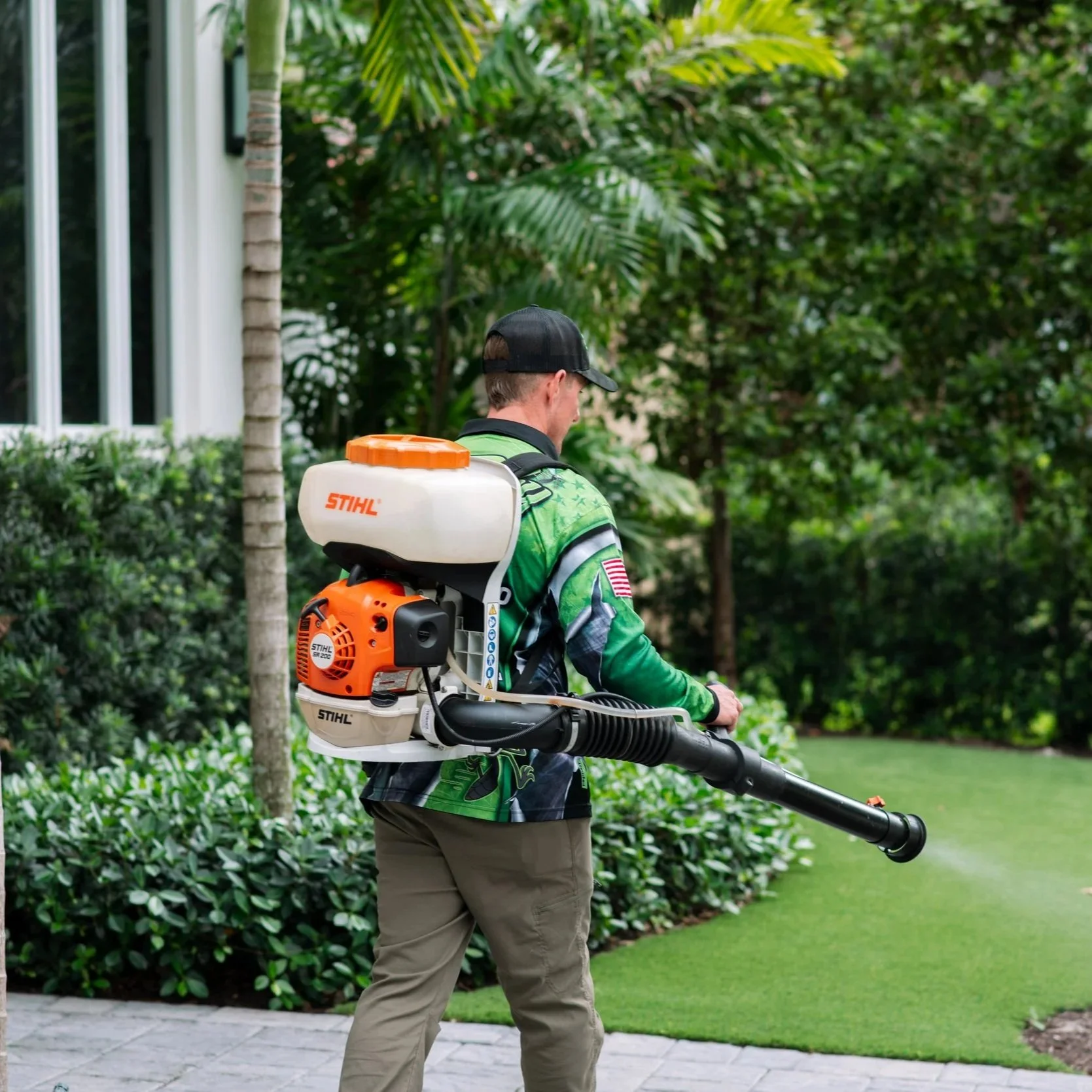 Man using a leaf blower outdoors in a yard with green bushes and trees, wearing a green shirt and a black cap.