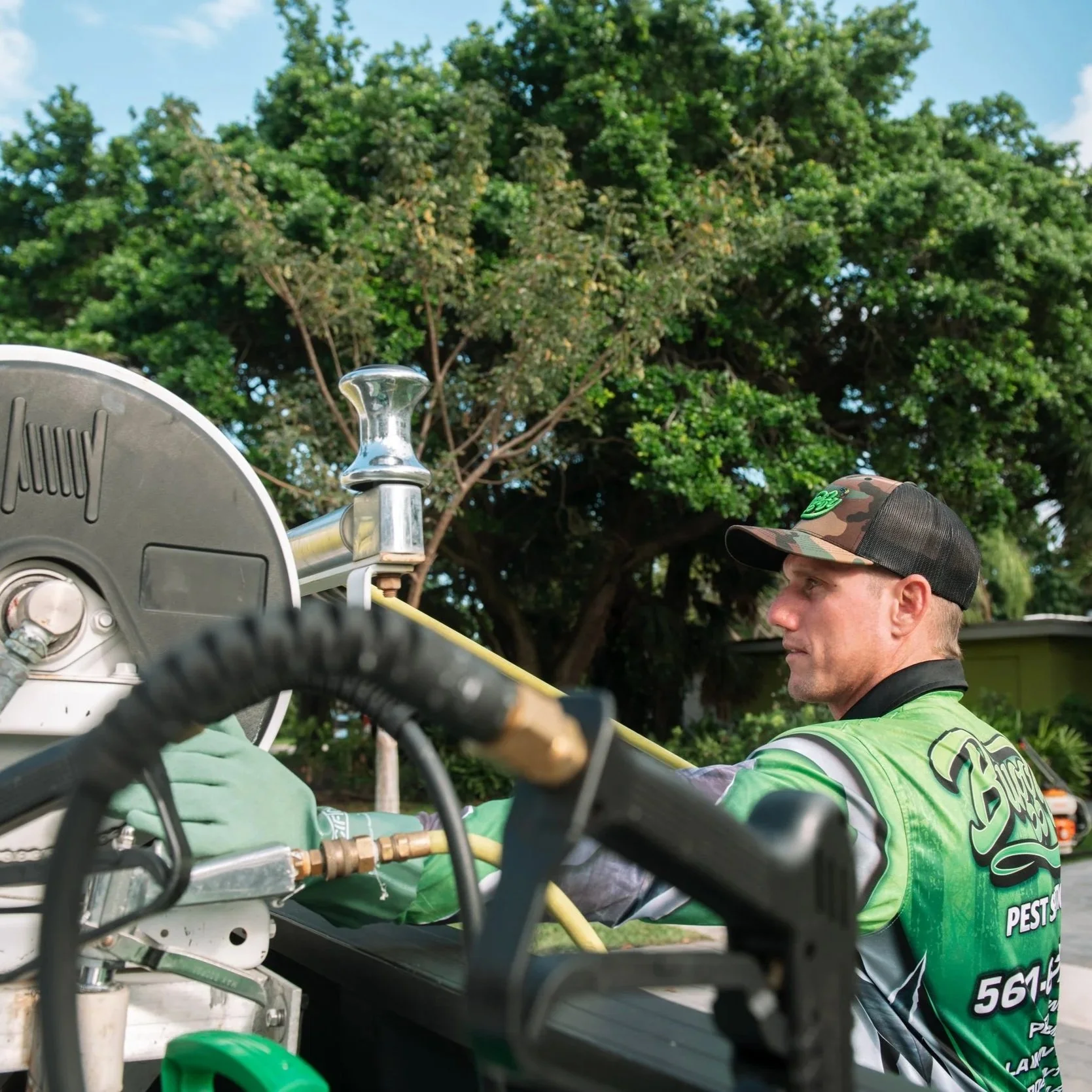 A man wearing a green and black shirt and a camo baseball cap working on equipment outdoors with green trees and blue sky in the background.