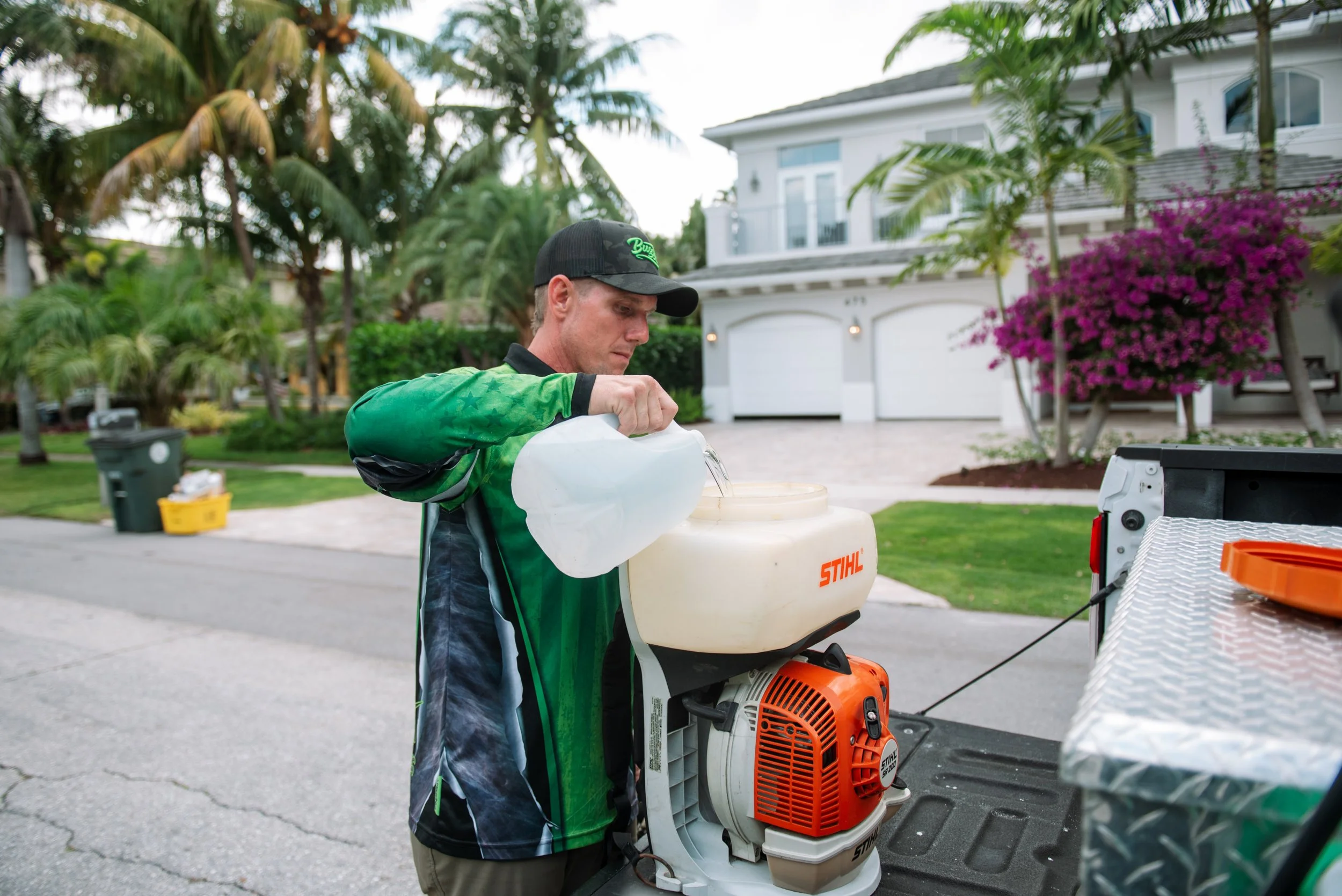 A man wearing a black cap and green and black shirt is filling a leaf blower with fuel from a white container. The scene takes place outdoors in a residential neighborhood with palm trees, flowering bushes, and a white house in the background.